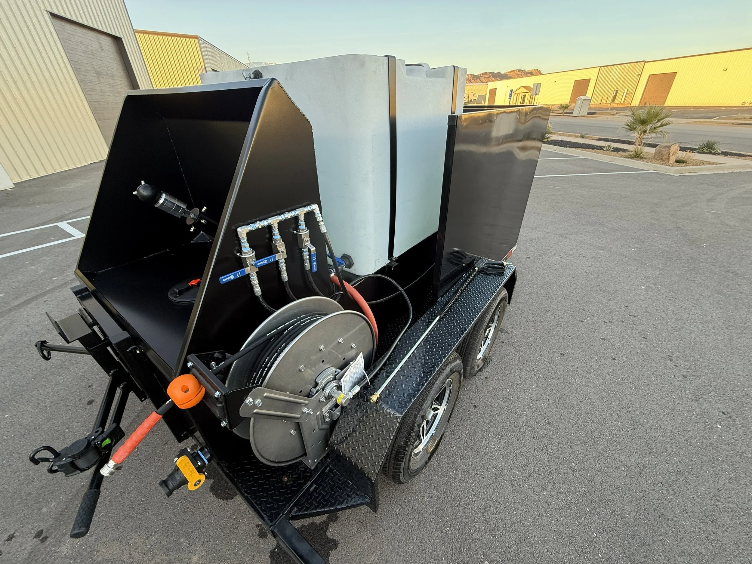A portable water or chemical spraying unit on a trailer, equipped with hoses, a reel, and a large white tank, situated in an empty parking lot with industrial buildings in the background.