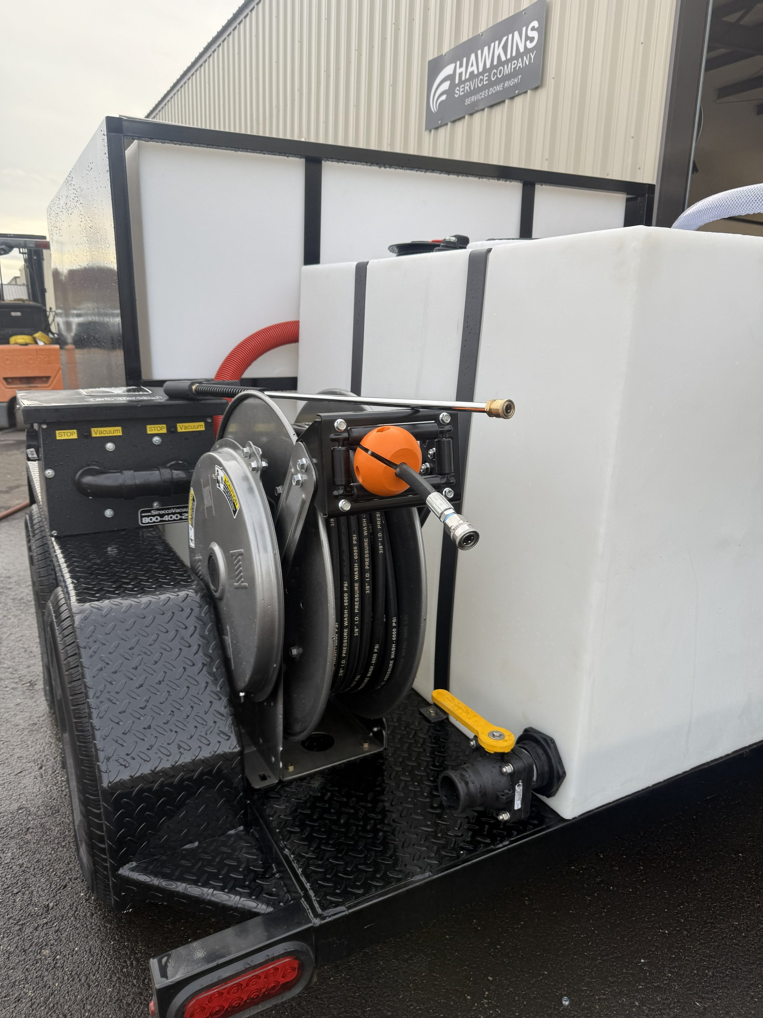 Close-up of a mobile water tank system with hoses, a reel, and a pump on a trailer. In the background, there is a building with a sign that reads 'Hawkins Service Company.'