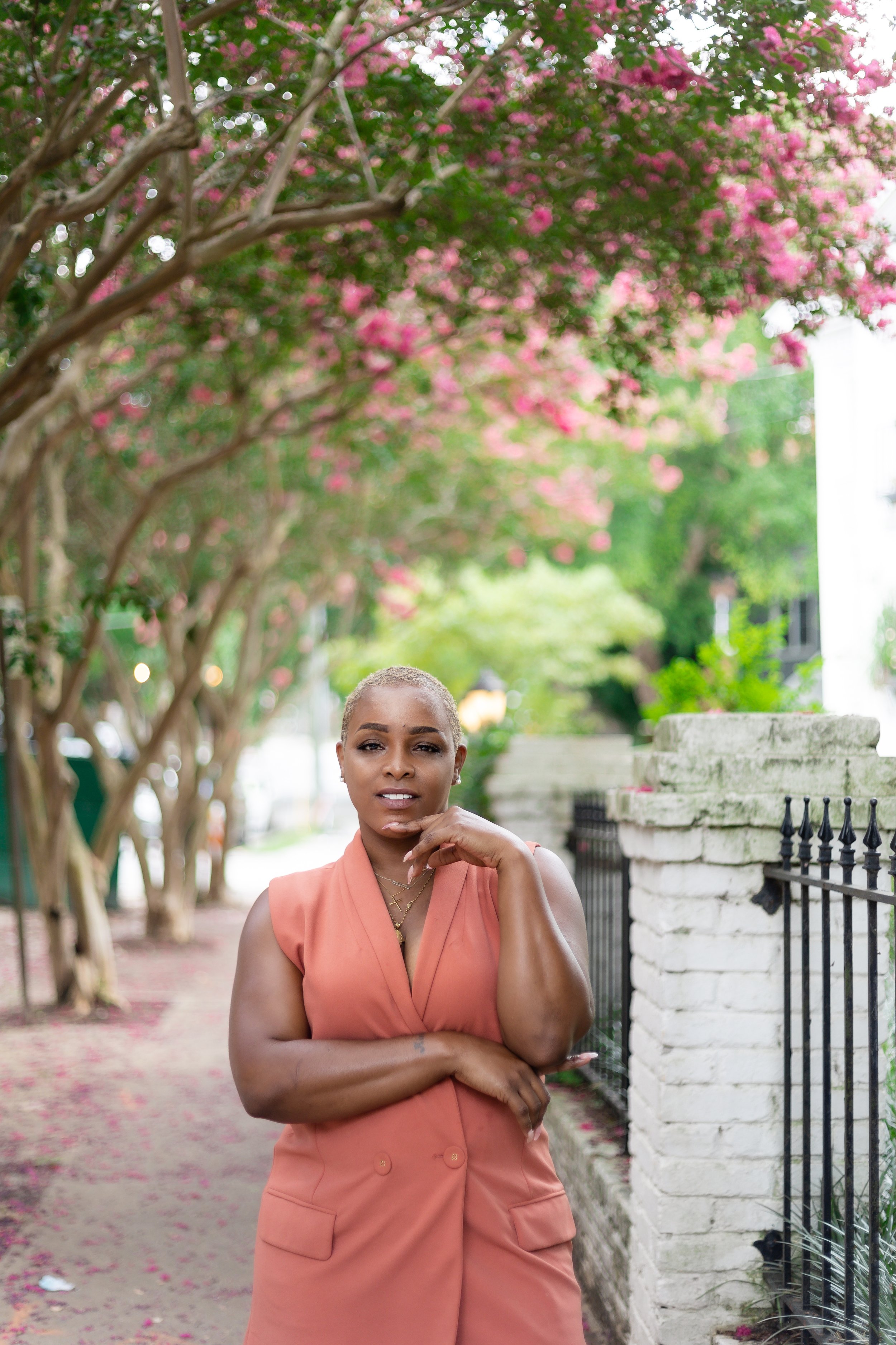 brown skin woman with short blonde haircut wearing a salmon blazer dress with her left arm folded in front of her waist, right elbow propped up with her chin resting against her hand