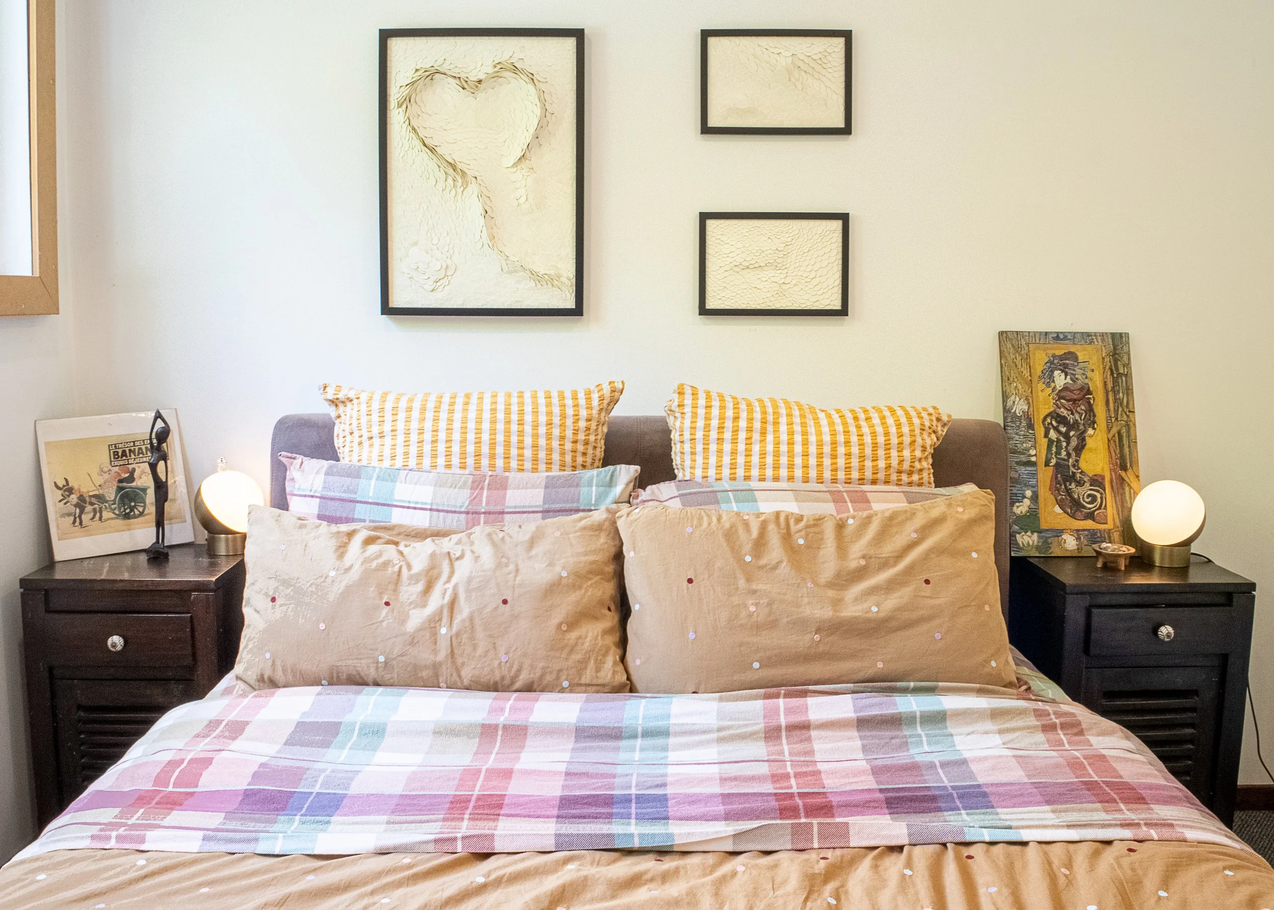 A bedroom with a neatly made bed featuring a plaid multicolored blanket and pillows with yellow and white striped pillowcases. Two dark wooden nightstands are on each side of the bed, with a framed picture of a vintage chariot and animals on the left and another framed piece of artwork on the right. A small lamp with a round white shade is on each nightstand.