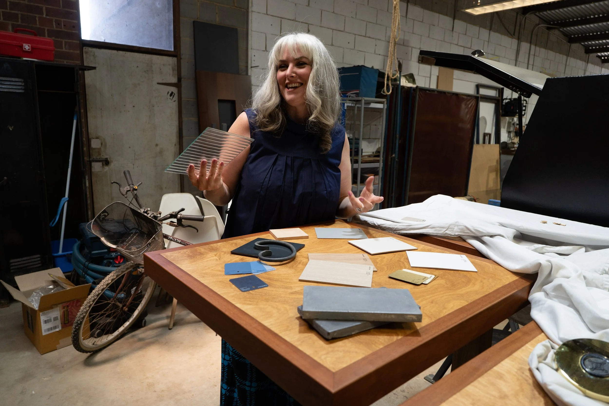 Woman with gray hair smiling and holding a glass object in a workshop with various tools, fabrics, and a bicycle nearby.
