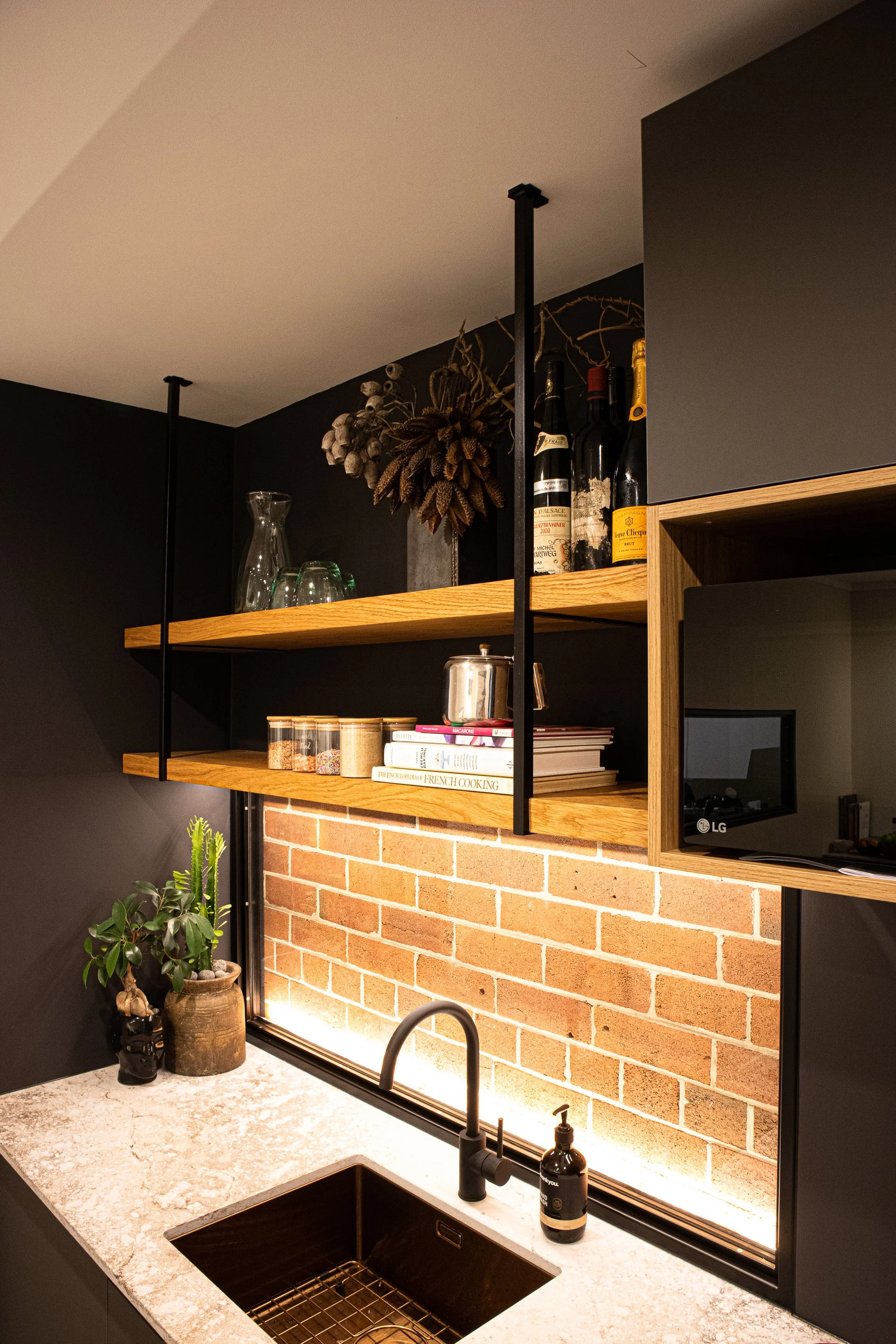 Modern kitchen with black walls, white marble countertop, black sink, black faucet, brick backsplash, open wooden shelves with wine bottles, glasses, and decorative plant, and black pendant lights.