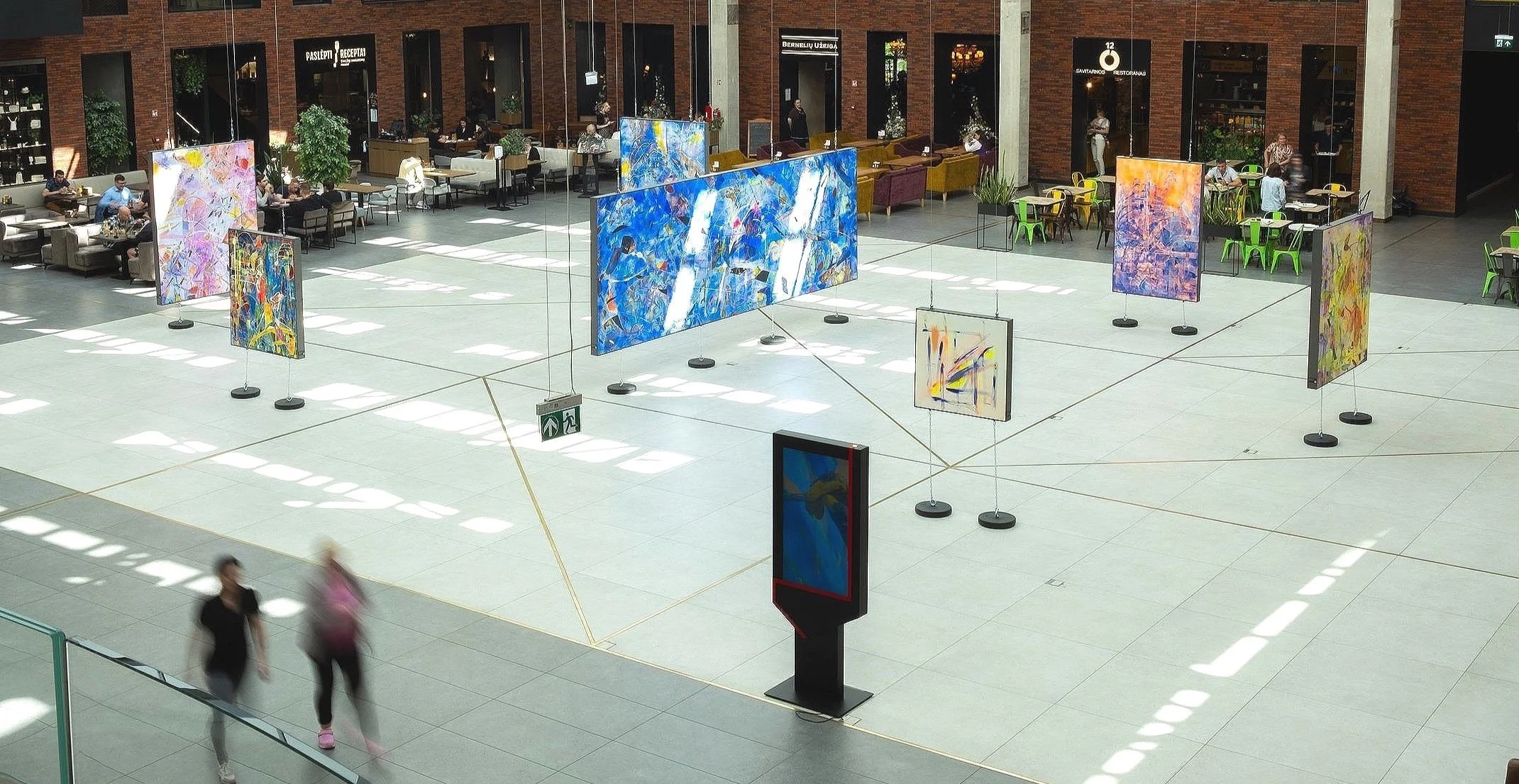 Art exhibition in a spacious indoor mall with colorful abstract paintings displayed on hanging panels and walls. Some people are walking by, and there are cafes and shops in the background.