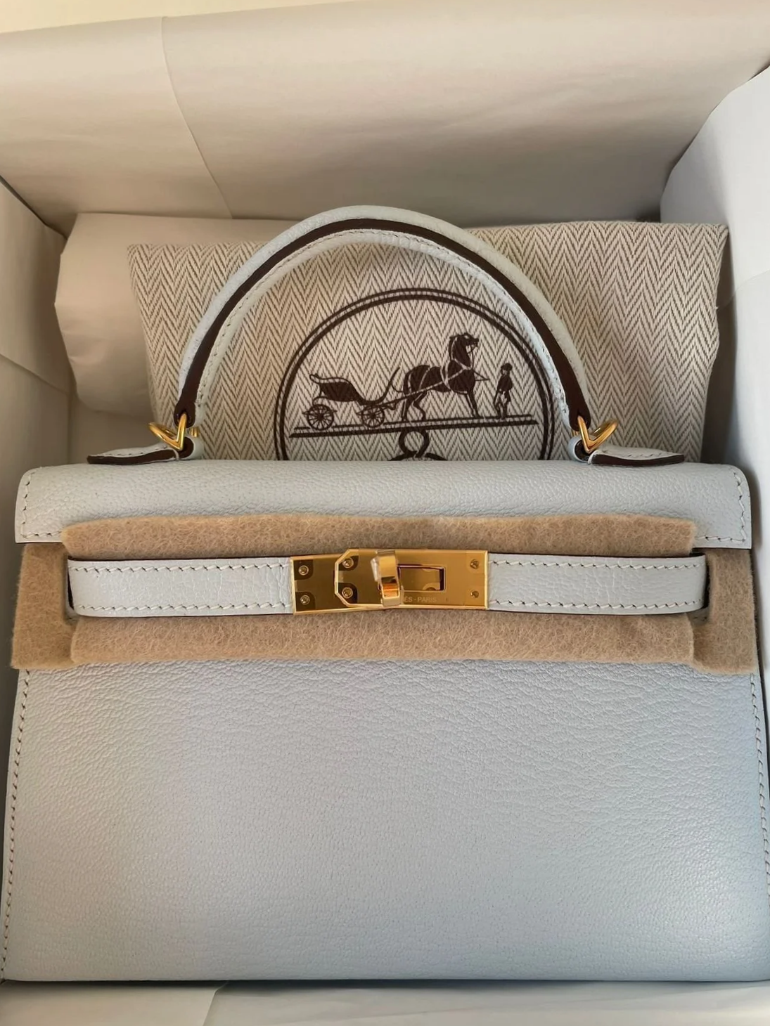 A white luxury handbag on top of a beige felt pad, placed on a textured beige surface. Behind the bag, there is a pillow with a design featuring a horse pulling a carriage with a person standing nearby.