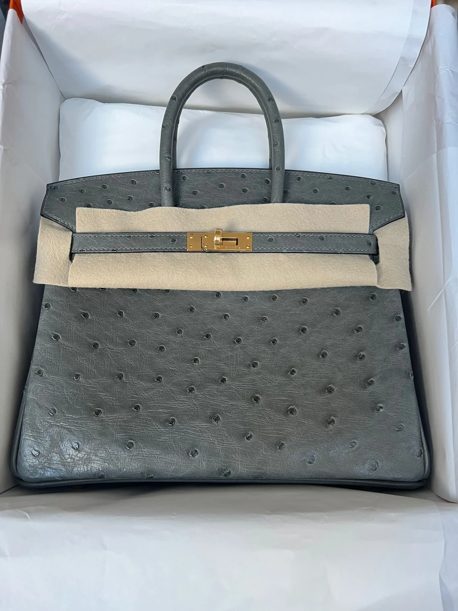 A gray handbag with ostrich leather texture and gold hardware, partially wrapped in protective felt, inside a white box.