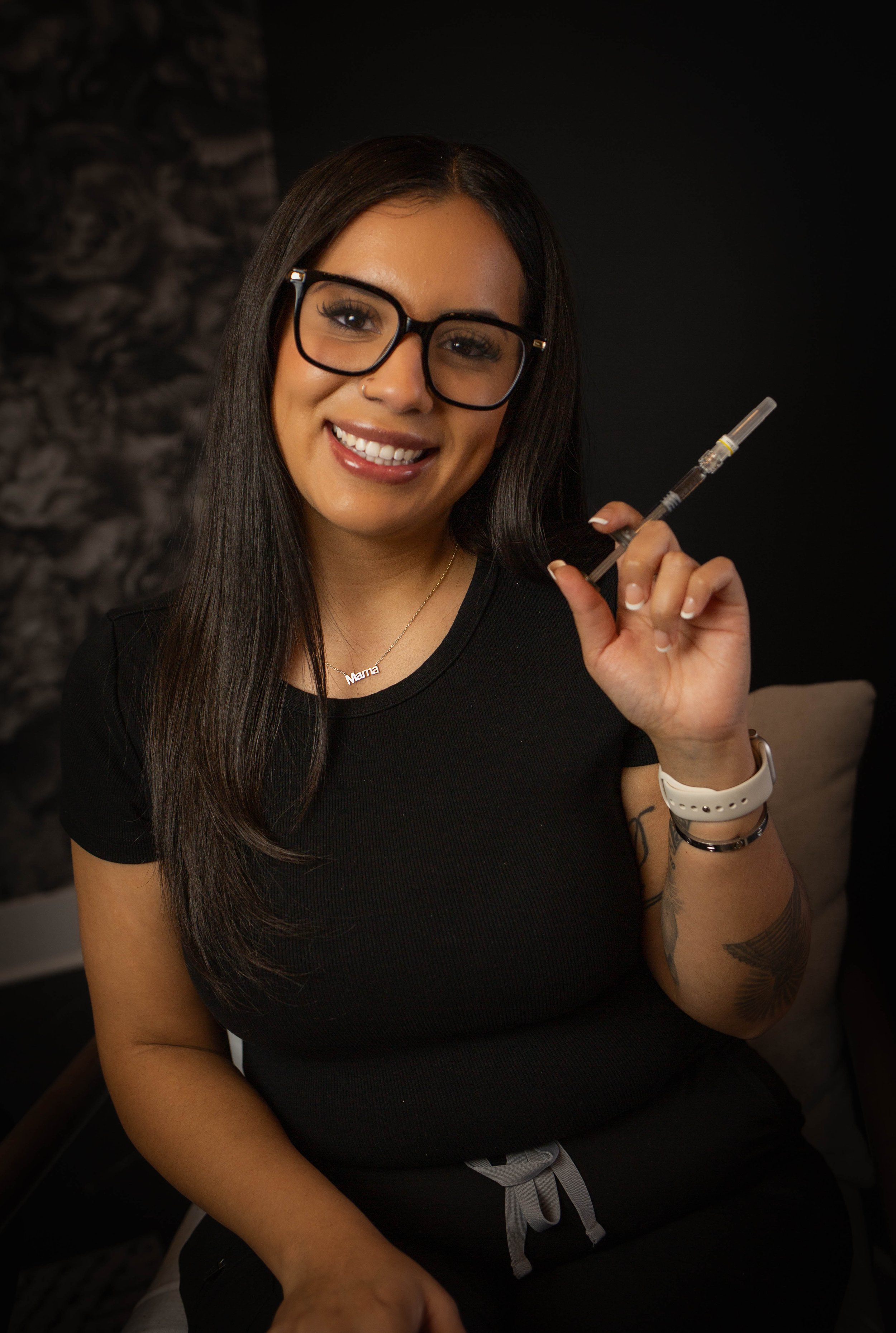 A woman with long black hair, wearing clear glasses, a black top, and a gold necklace with a name pendant, smiling in front of a gray textured background.