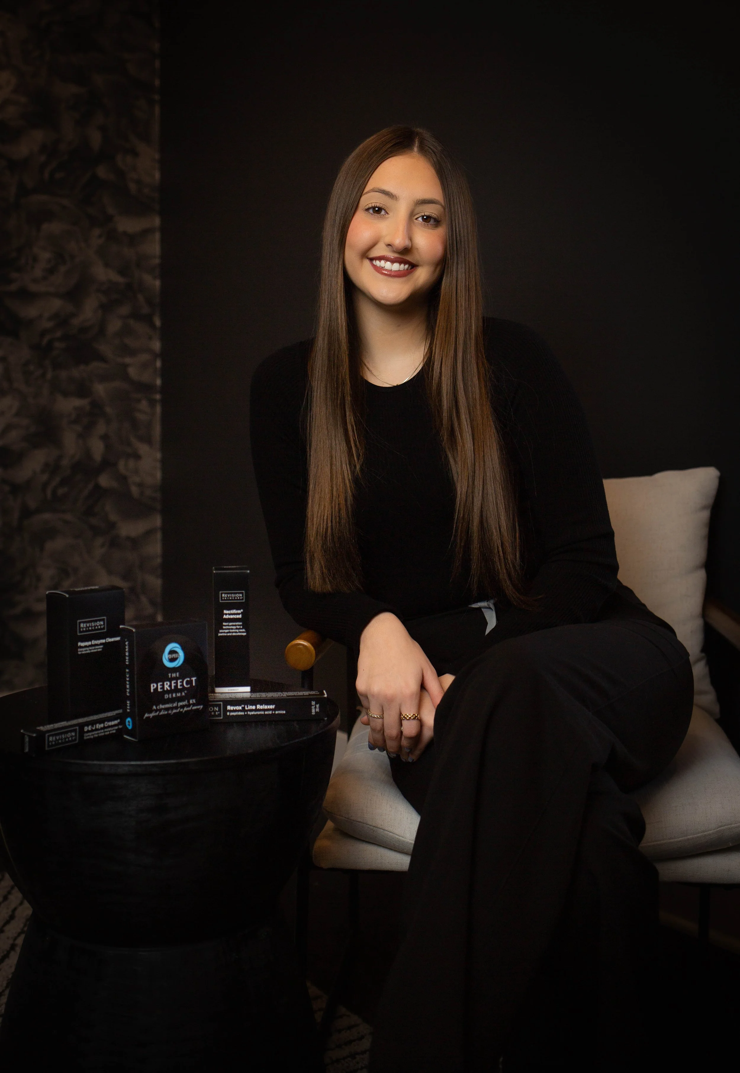 A young woman with long brown hair smiling in front of a floral patterned wall.