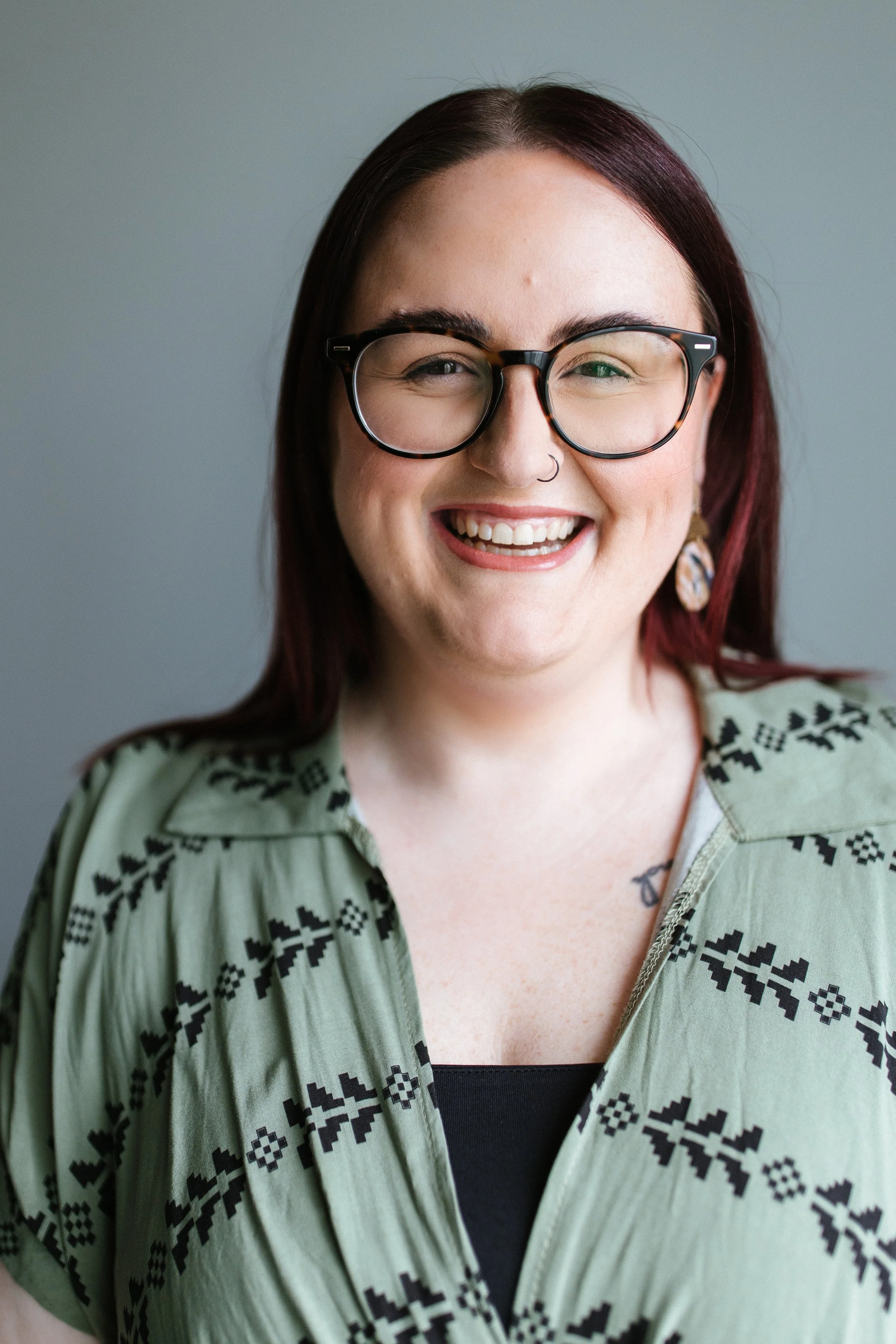 A woman with dark red hair, wearing glasses, smiling, and dressed in a green patterned shirt over a black top.