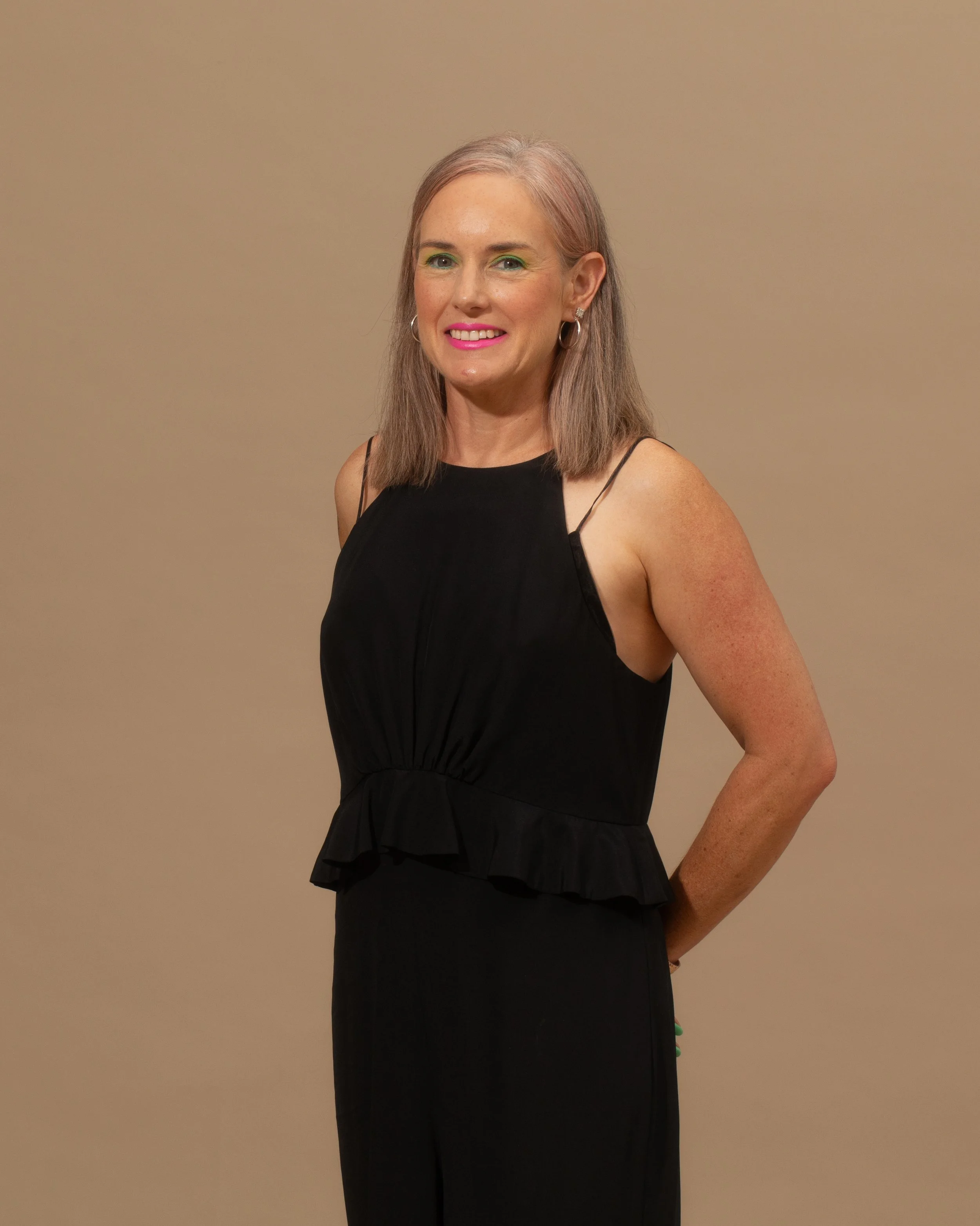 A woman with shoulder-length light brown hair, wearing a black sleeveless dress, standing against a plain beige background, smiling with pink lipstick and silver hoop earrings. Laura McConnell Conti