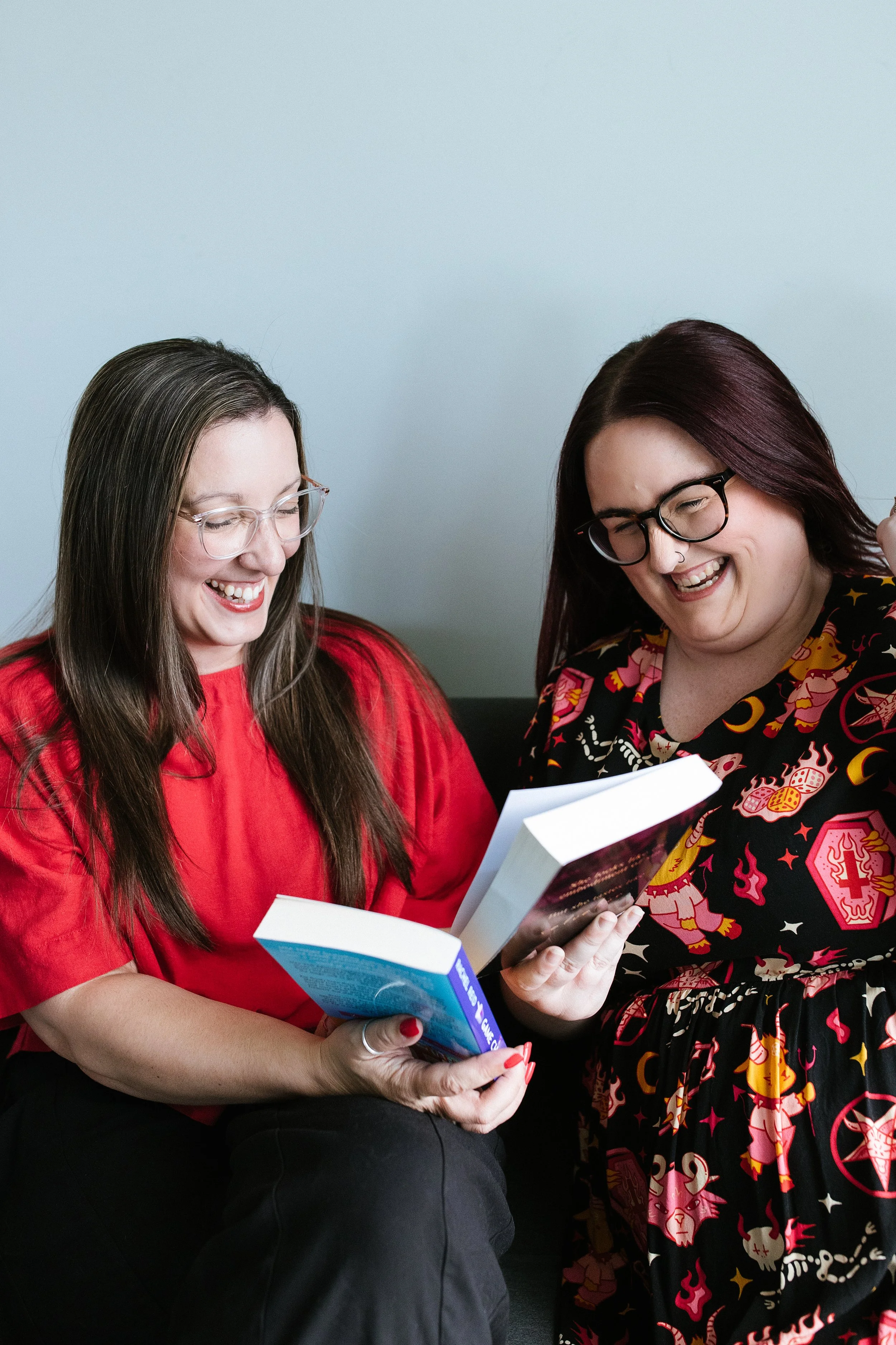 Two women sitting together, smiling and reading books, Elise Heerde wearing a red top with long brown hair and glasses, Sam Sellers wearing a black shirt with a colorful tattoo-style pattern, with purple hair and glasses.