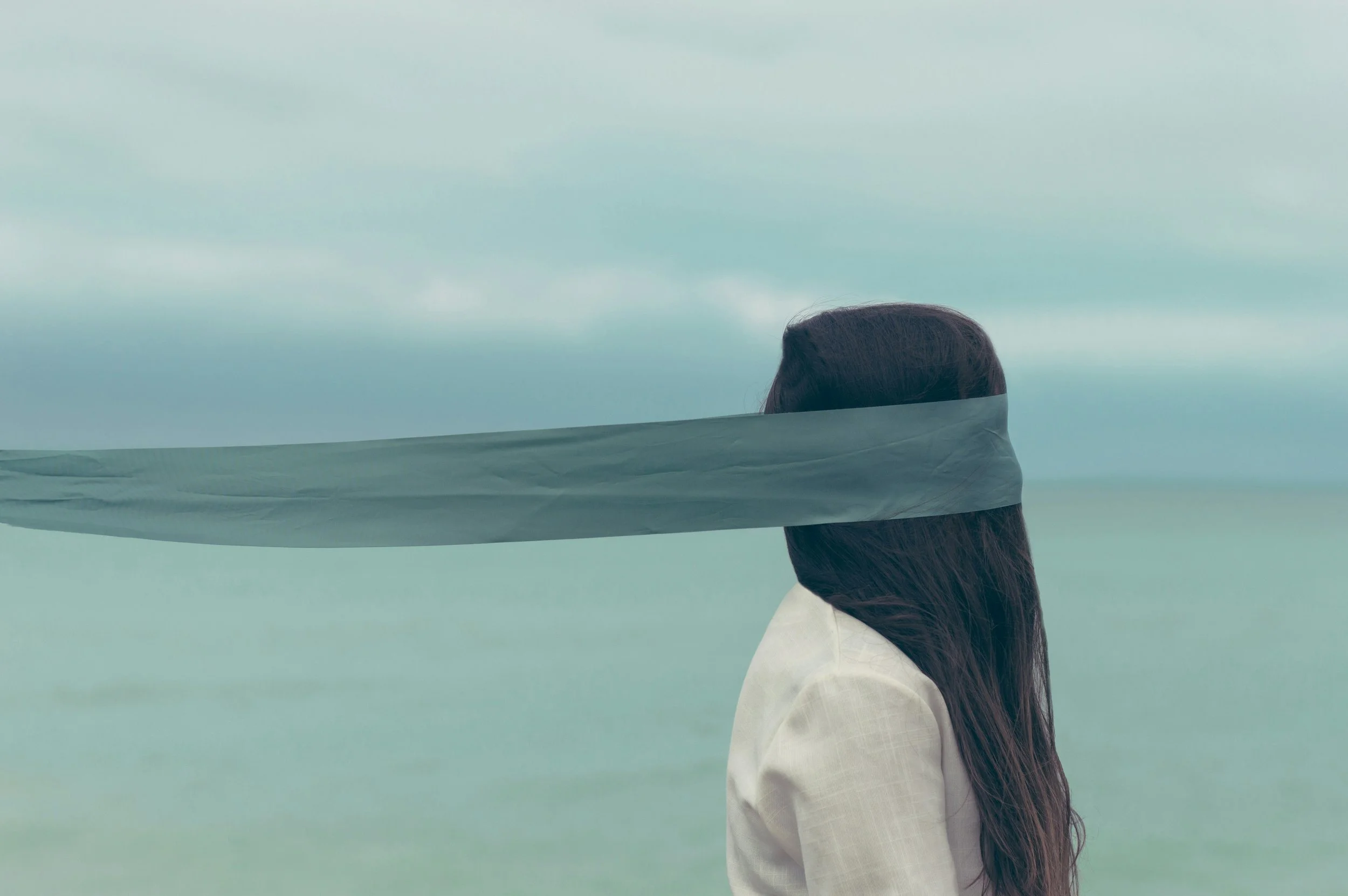 Person with long dark hair facing sideways in front of a cloudy sky, with a grey ribbon or cloth across their face.