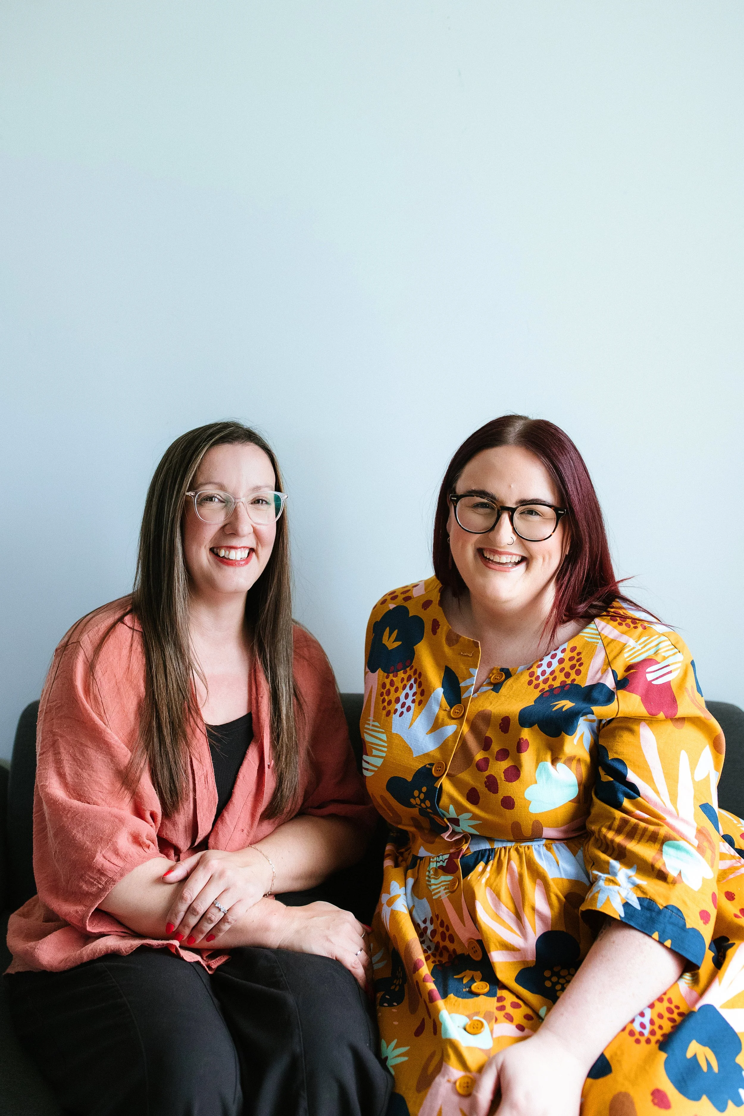Two women sitting together and smiling at the camera against a light blue wall, one wearing glasses and a floral yellow dress, the other wearing glasses and a pinkish-brown cardigan.