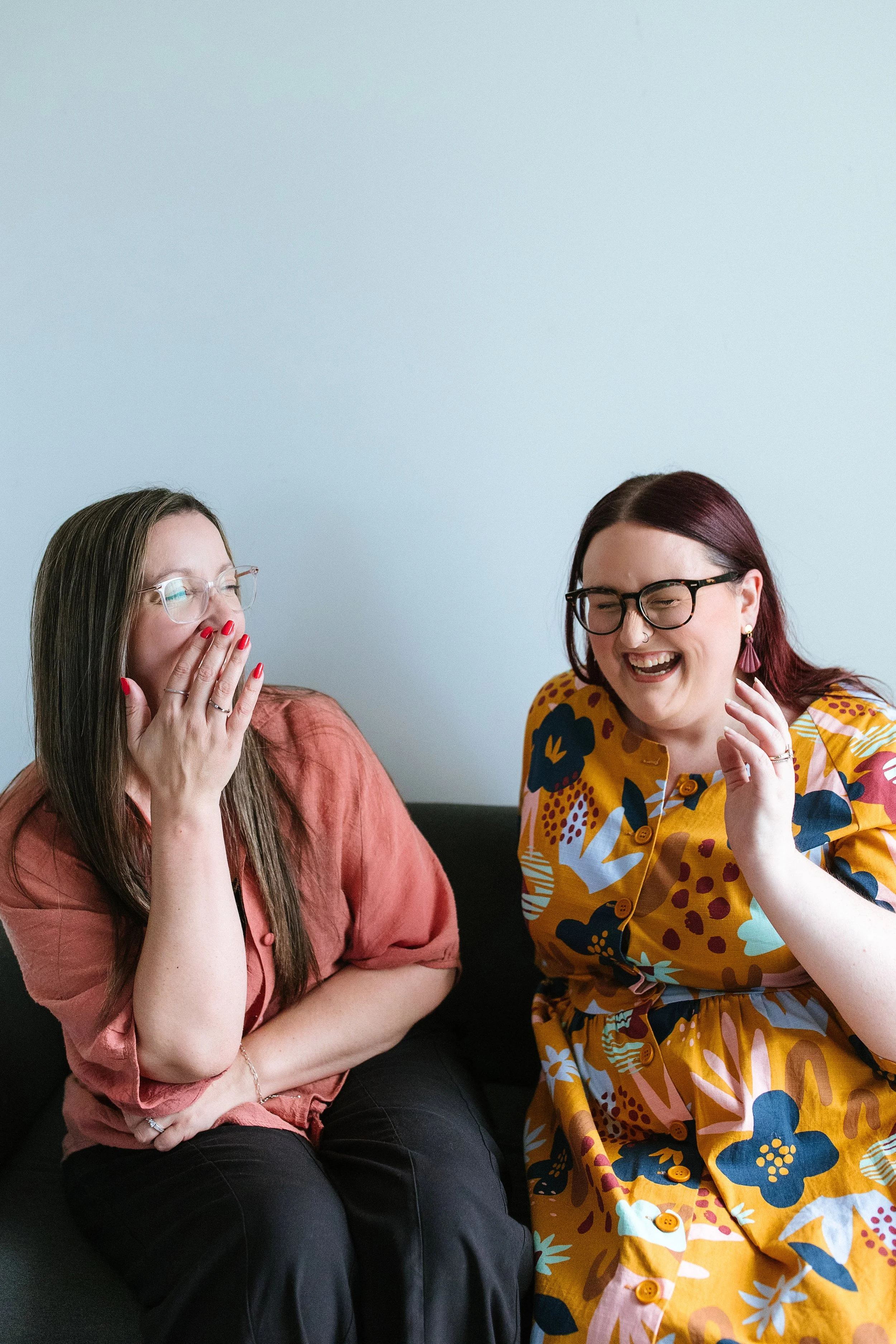 Two women sitting close together on a black couch against a light blue wall, sharing a laugh. The woman on the left has long brown hair, glasses, and is wearing a pinkish-brown top with red nail polish. The woman on the right has dark red hair, glasses, and is wearing a colorful, patterned yellow dress with large floral and abstract designs.