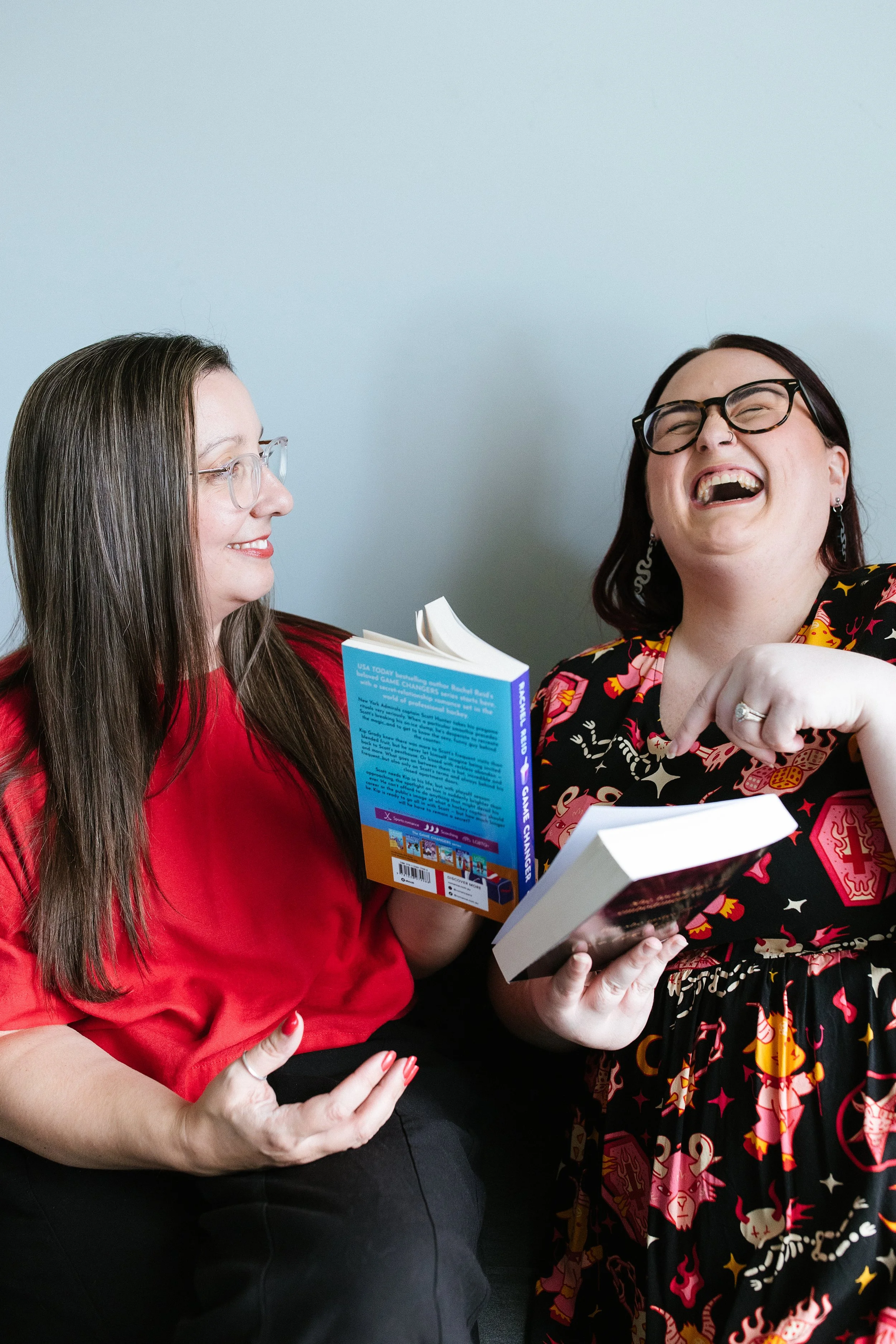 Two women sitting close together, smiling and laughing while reading and sharing books in a casual setting. Elise Heerde & Sam Sellers