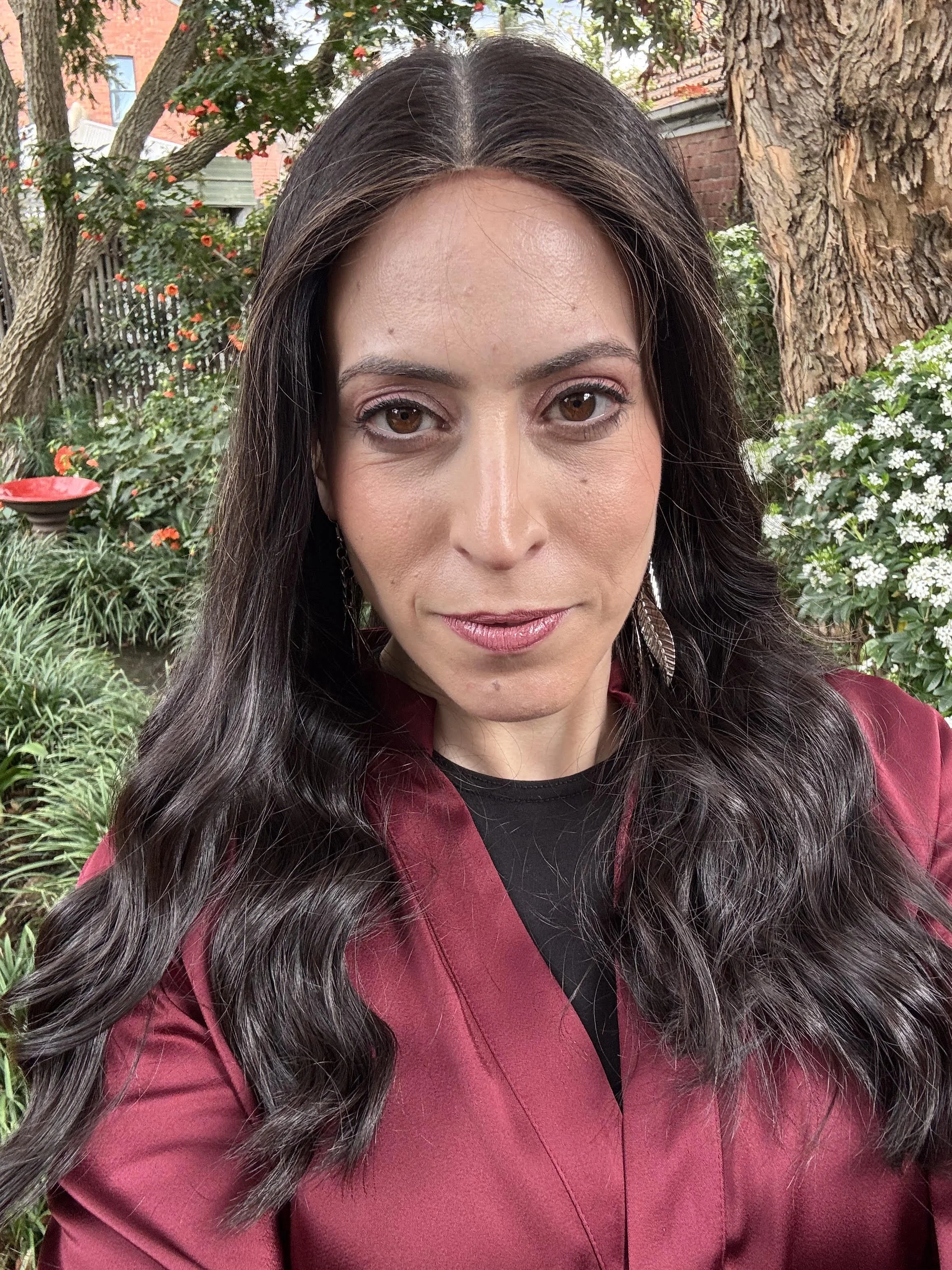 Close-up of a woman with long dark wavy hair, wearing a maroon blazer and earrings, outdoors with trees and plants in the background. Nicole Meyer