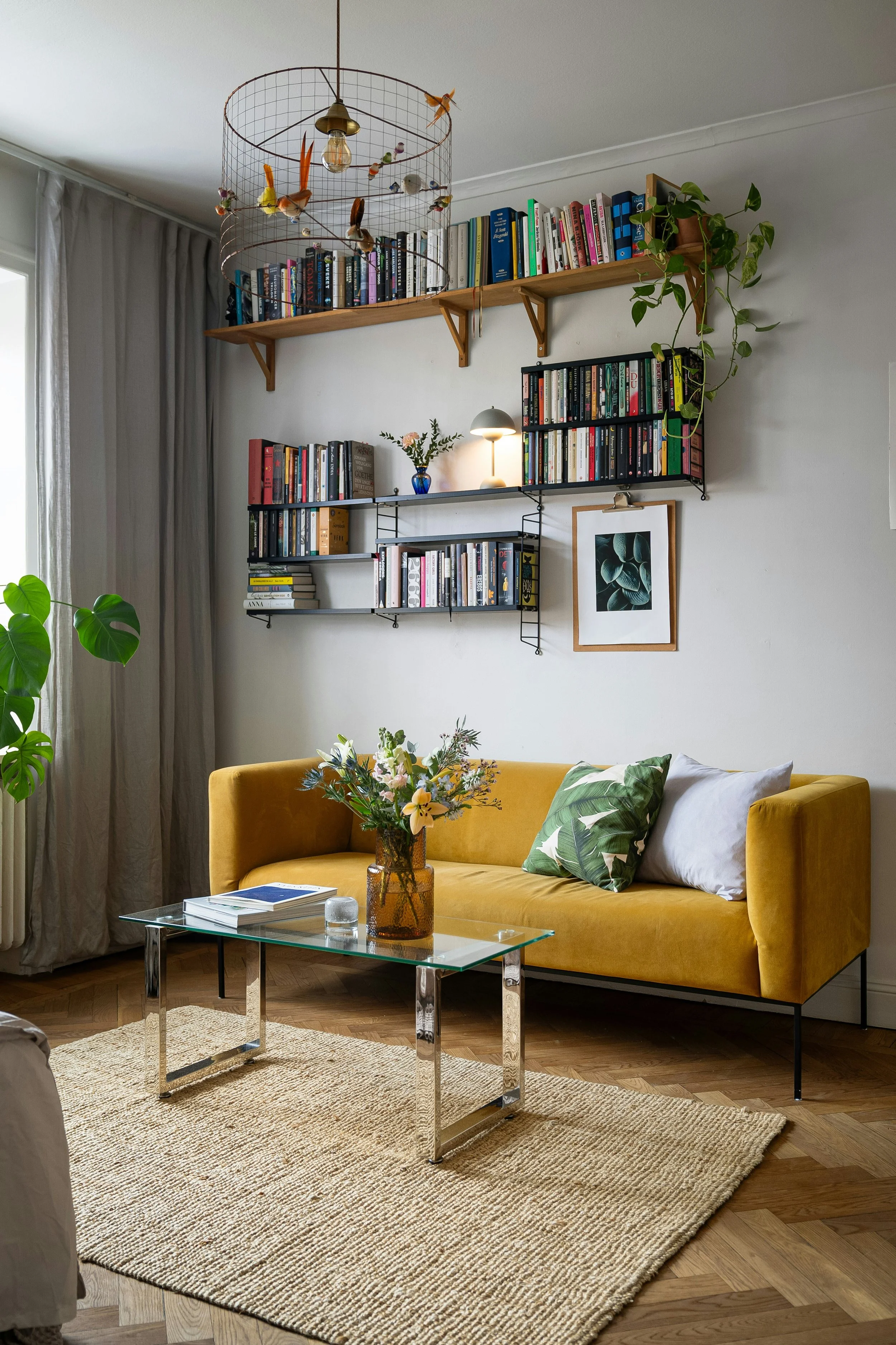 Living room with a yellow sofa, glass coffee table, potted plants, wall-mounted bookshelves, and a decorative light fixture.