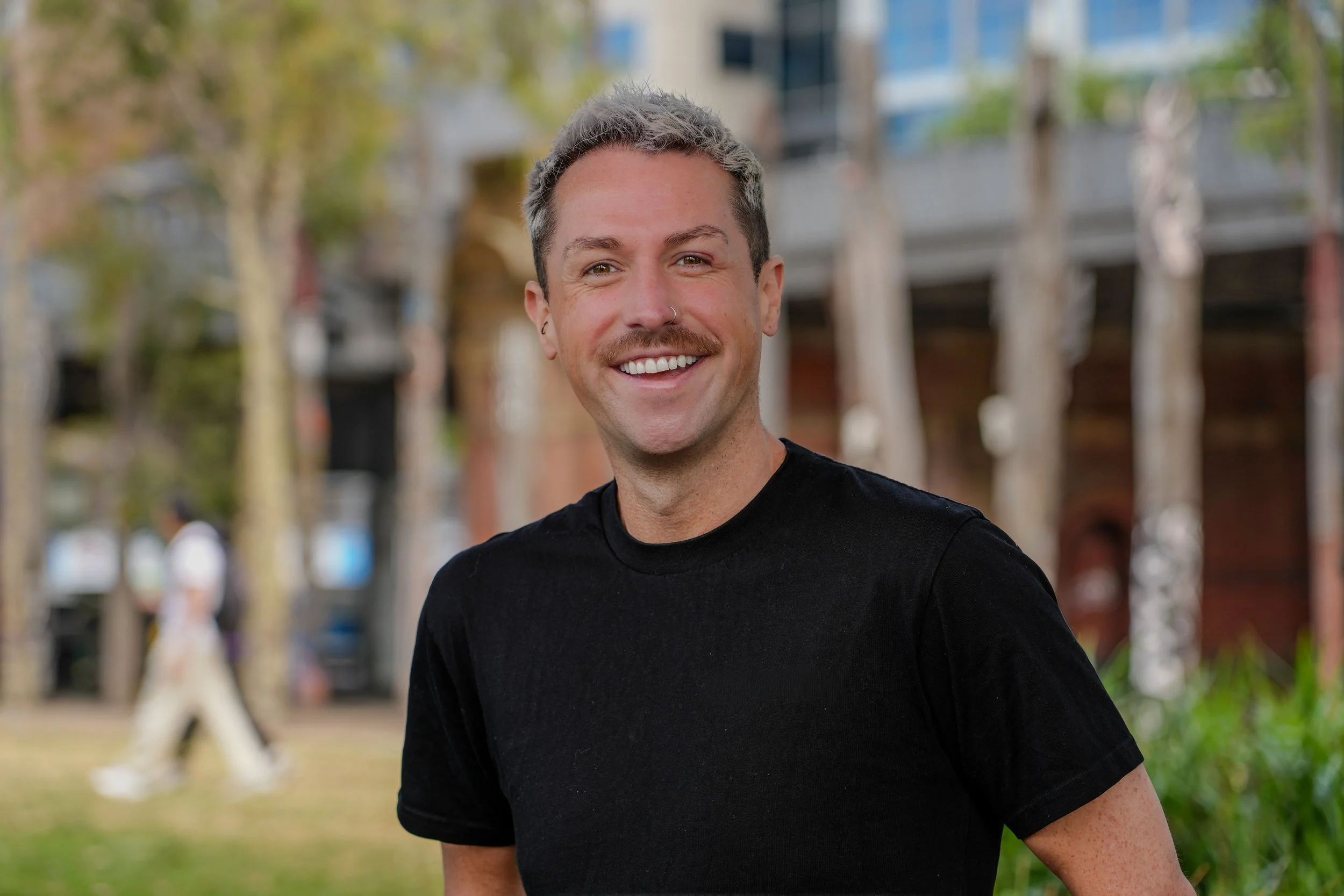 A smiling man with short gray hair, wearing a black shirt, outdoors in a park with trees and buildings in the background. Aaron Kelly