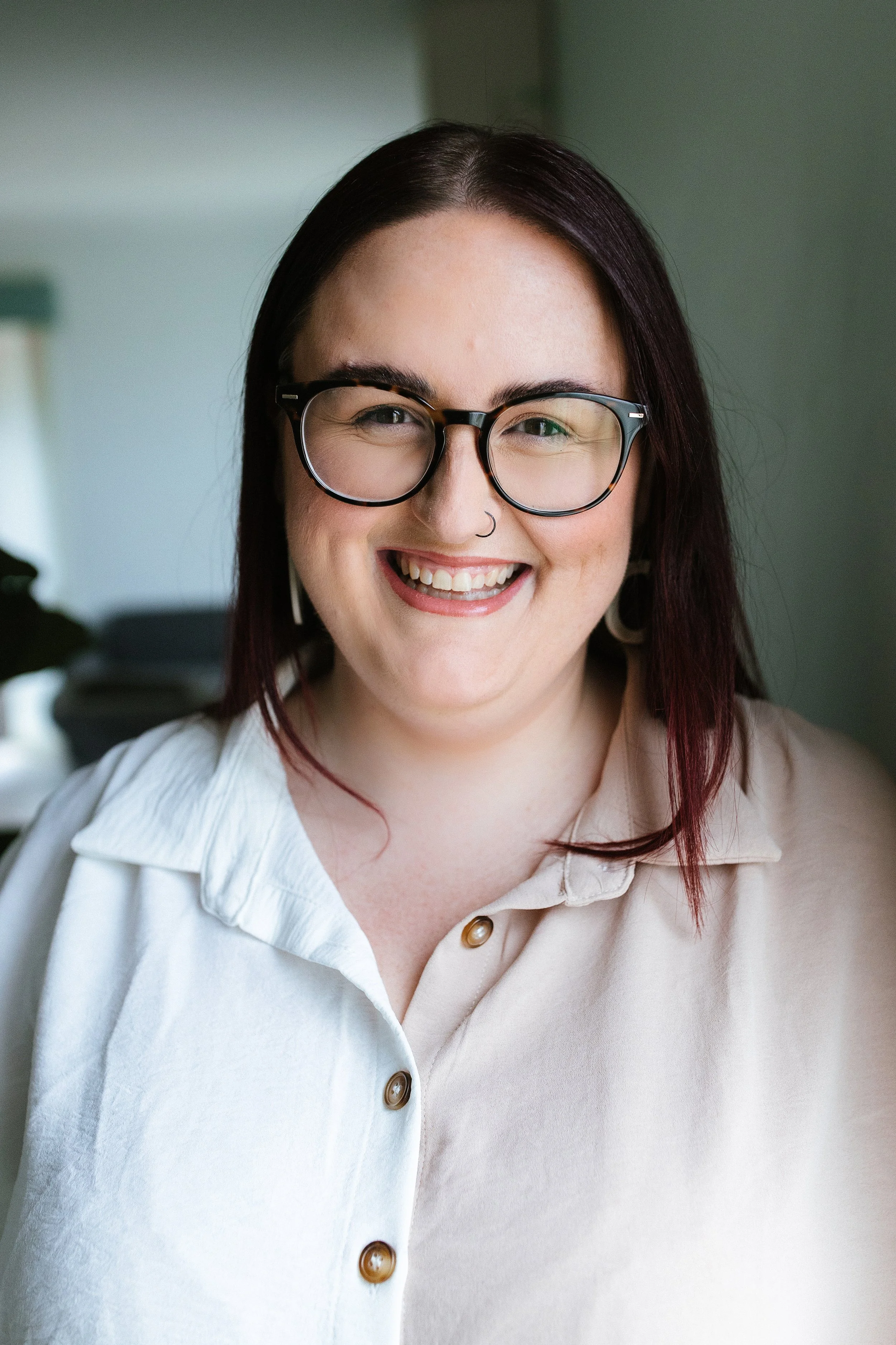 Close-up of a woman smiling, wearing black glasses, a nose ring, and earrings, with brunette hair with red tips, dressed in a cream-colored collared shirt. Sam Sellers