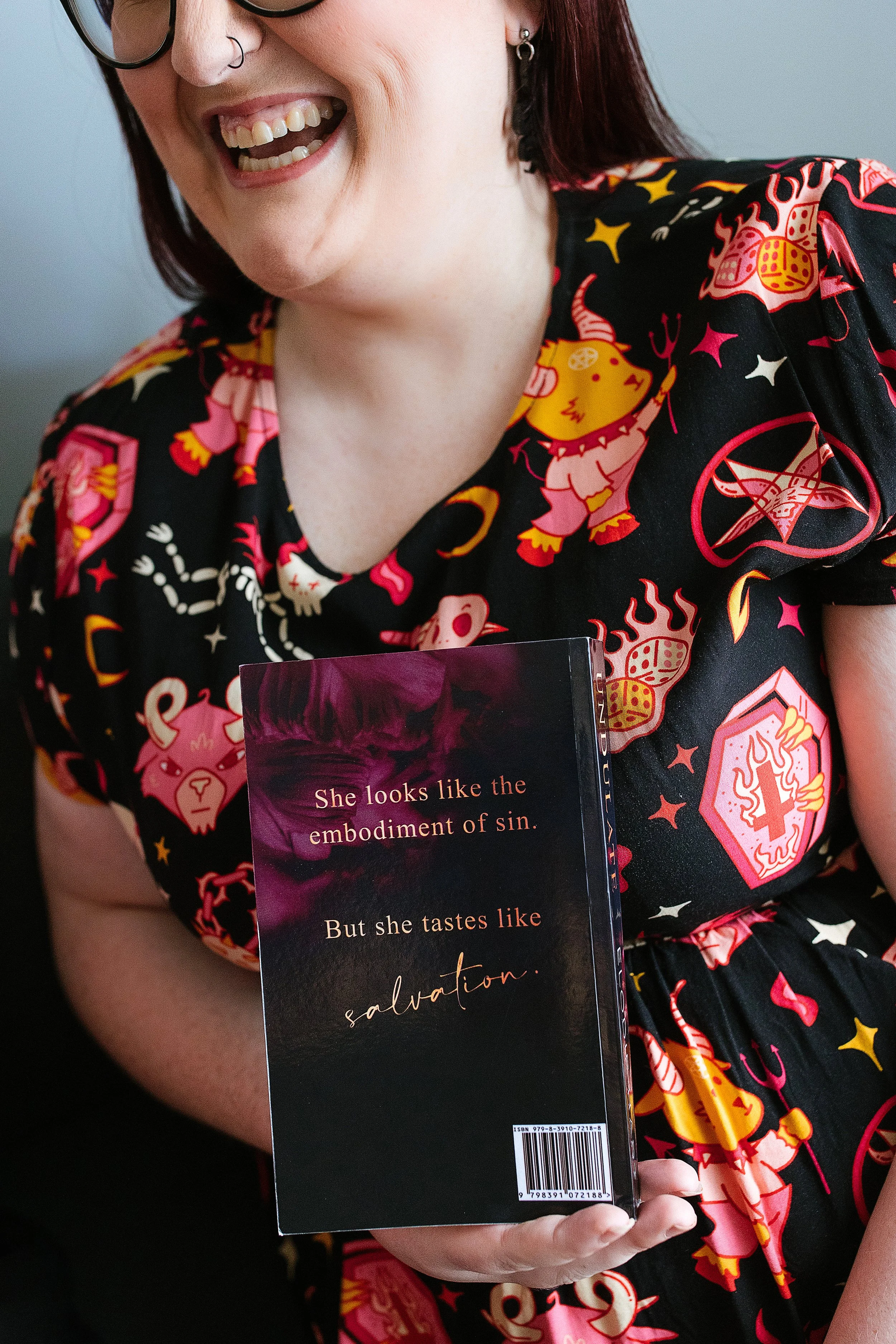 A person with glasses and piercings smiling while holding a book with a dark cover and text that reads, "She looks like the embodiment of sin. But she tastes like salvation." The person is wearing a black dress with colorful cartoon-style monsters and symbols.