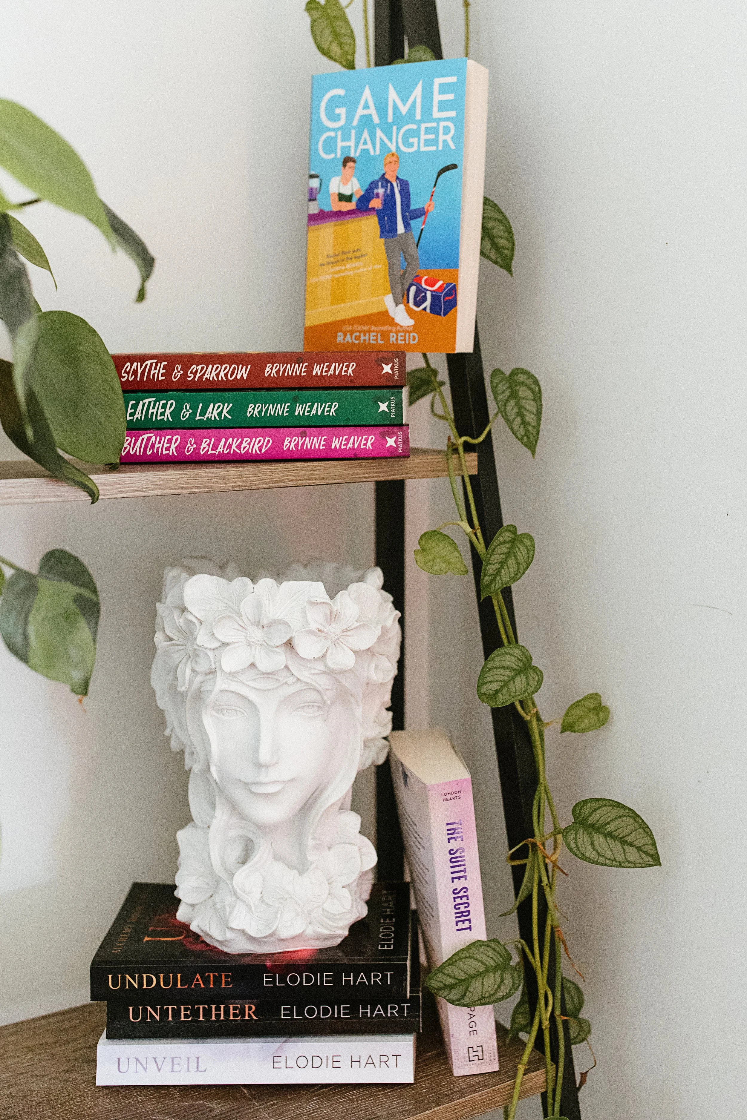 A white sculpture of a face with floral hair design sits on a table, with a stack of books titled 'Undulate,' 'Untether,' and 'Unveil' by Elodie Hart underneath it. A book titled 'Game Changer' by Rachel Reid is propped up on a black ladder shelf. Green vine plant with heart-shaped leaves drapes around the shelf.