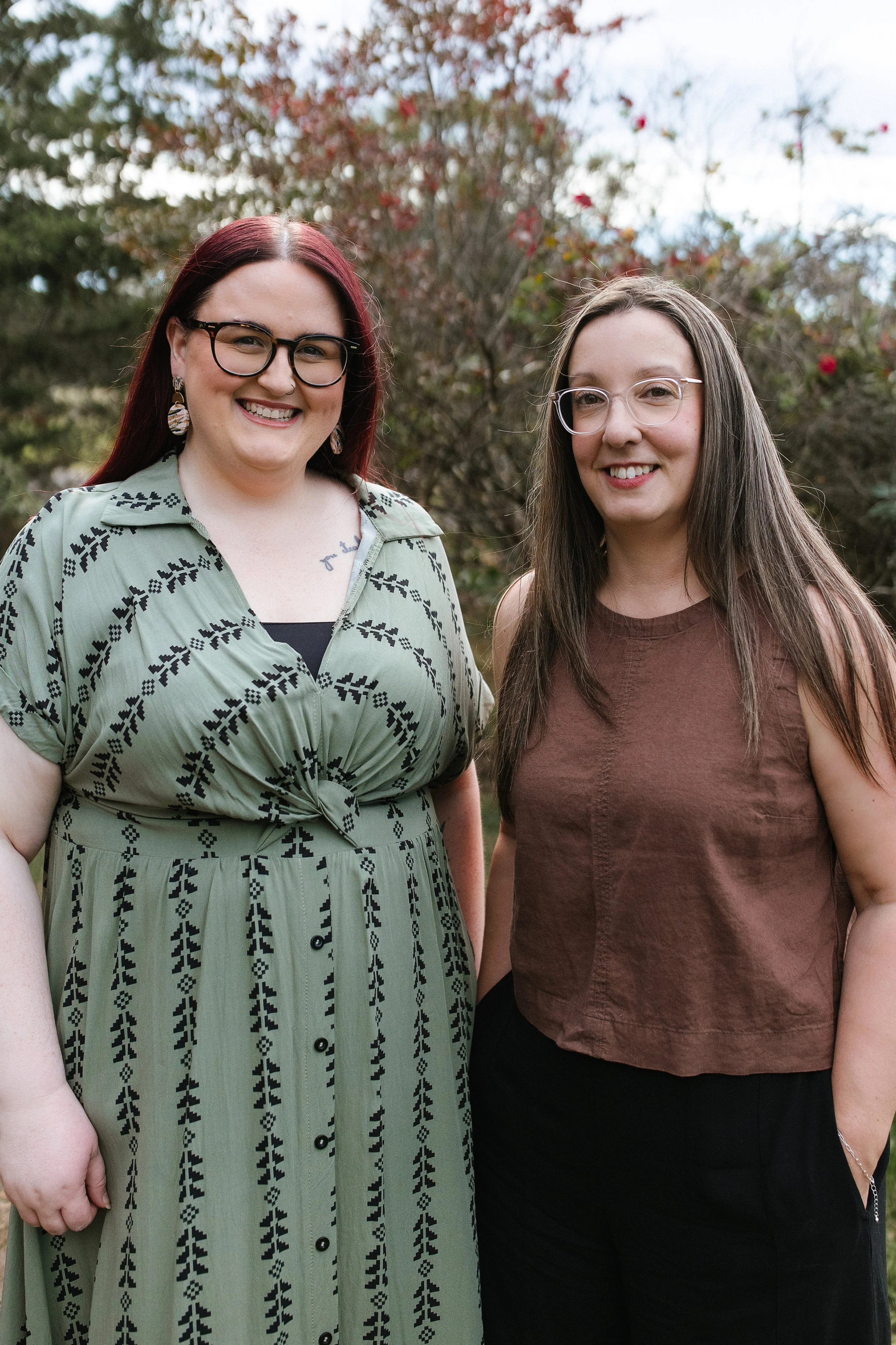 Two women standing outdoors in front of a shrub with red flowers, smiling at the camera. Elise Heerde & Sam Sellers