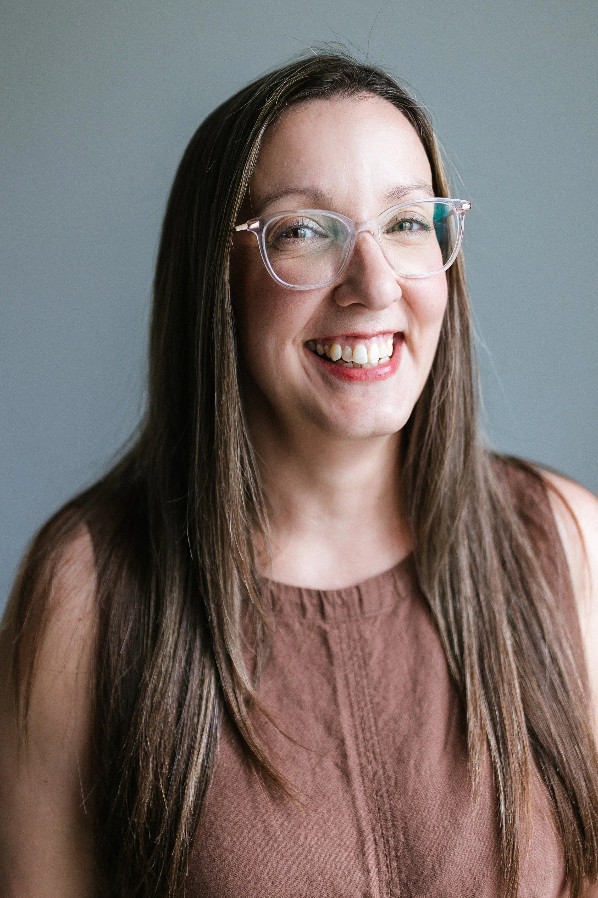 Close-up of a smiling woman with long, dark hair, wearing clear glasses and a brown sleeveless top, against a plain background.