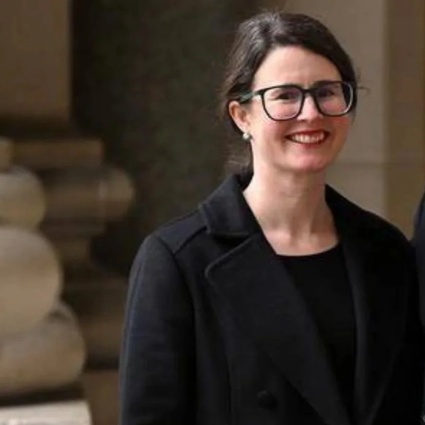 A woman with dark hair, glasses, and earrings, smiling and wearing a black coat indoors. Catherine Carey