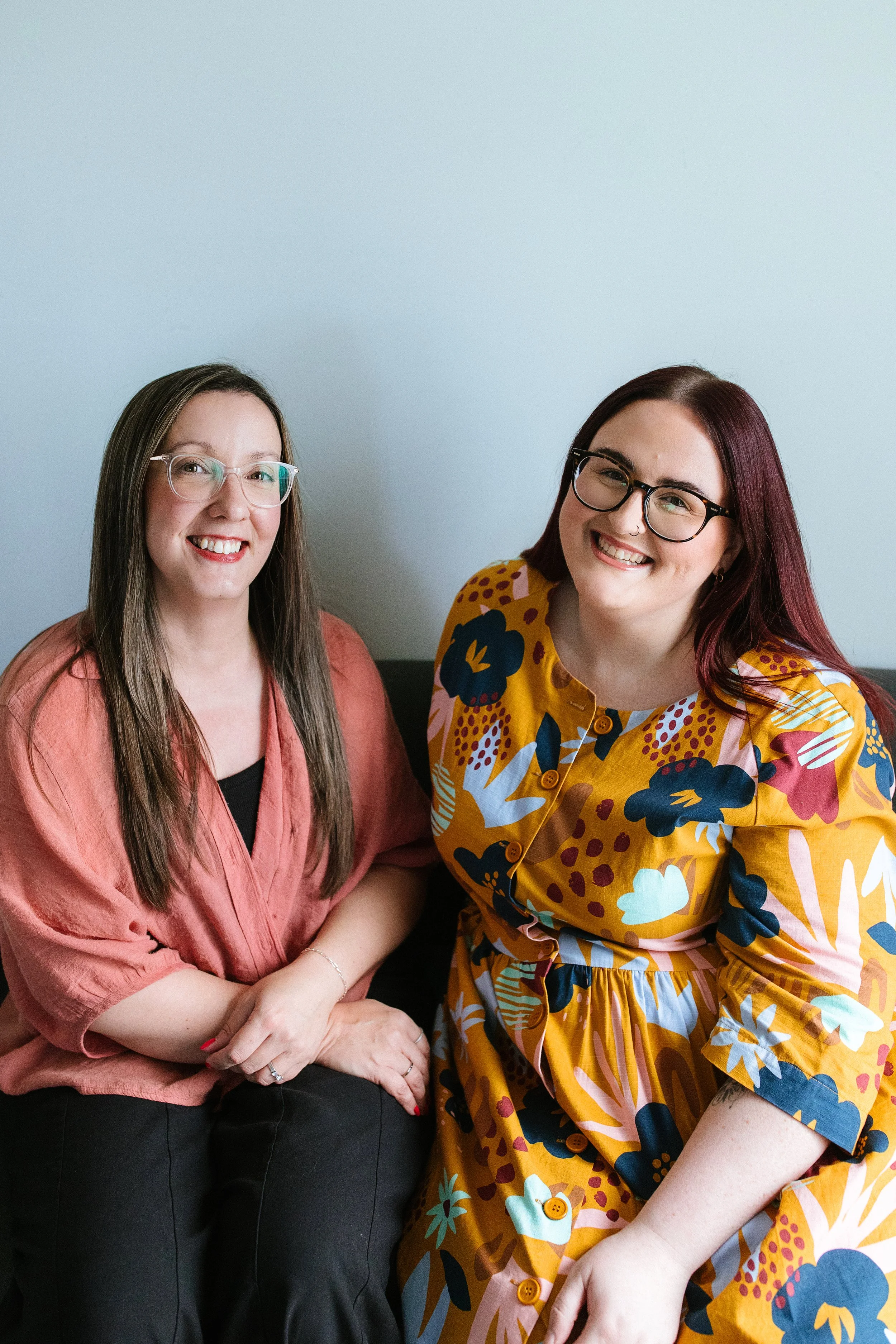 Two women sitting together smiling, one wearing glasses and a pink top, the other wearing glasses and a colorful floral-patterned dress. Elise Heerde & Sam Sellers