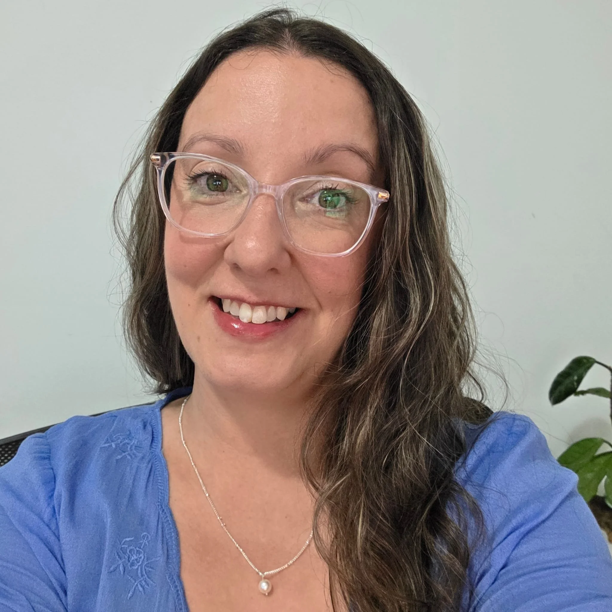 Woman with long hair wearing glasses and a brown sweater, standing in front of dense green foliage.