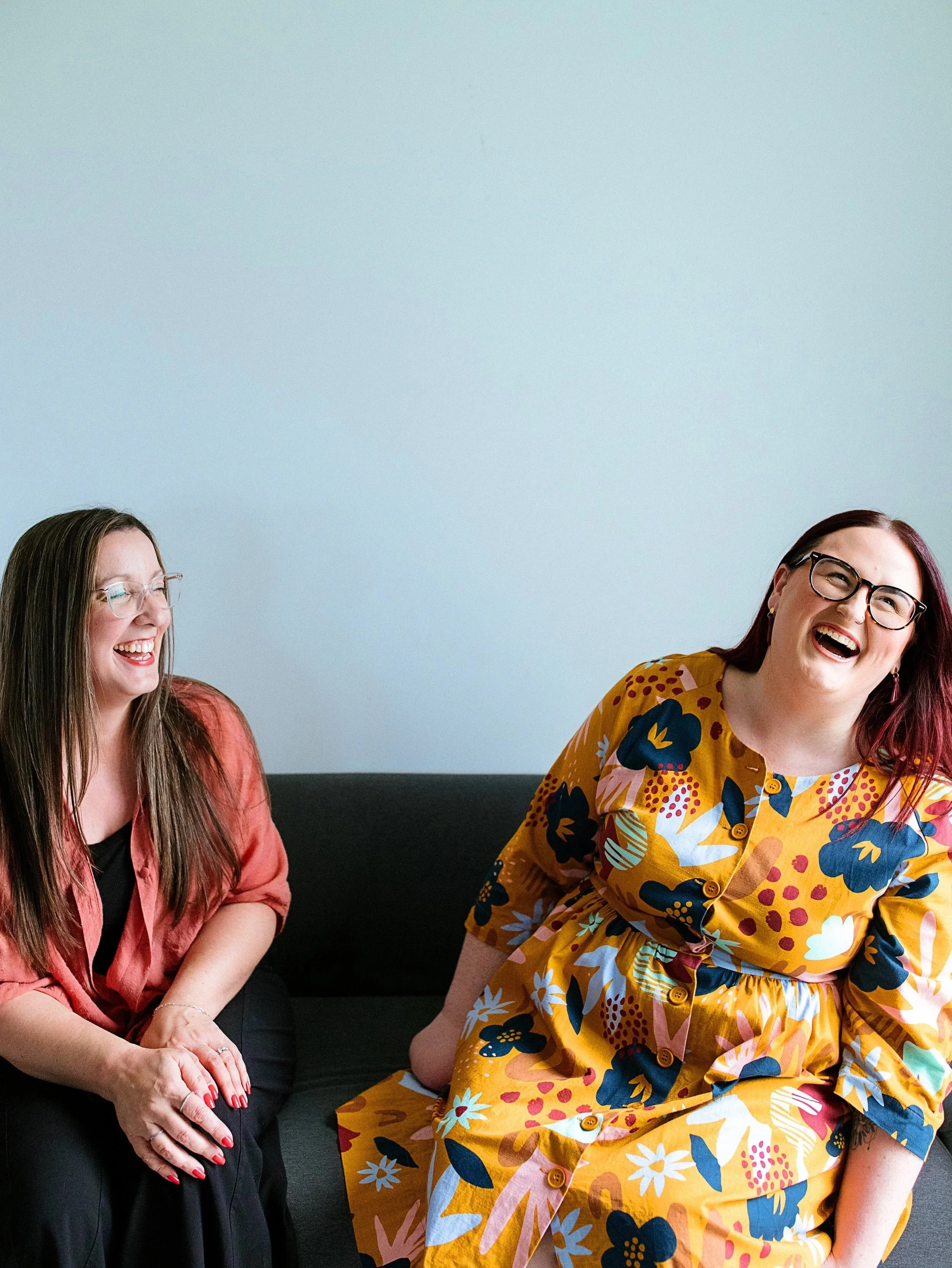 Two women sitting on a couch, laughing and smiling in a casual indoor setting. One woman has long brown hair and glasses, wearing an orange blazer. The other woman has long dark hair with red highlights, glasses, and is wearing a yellow floral dress.