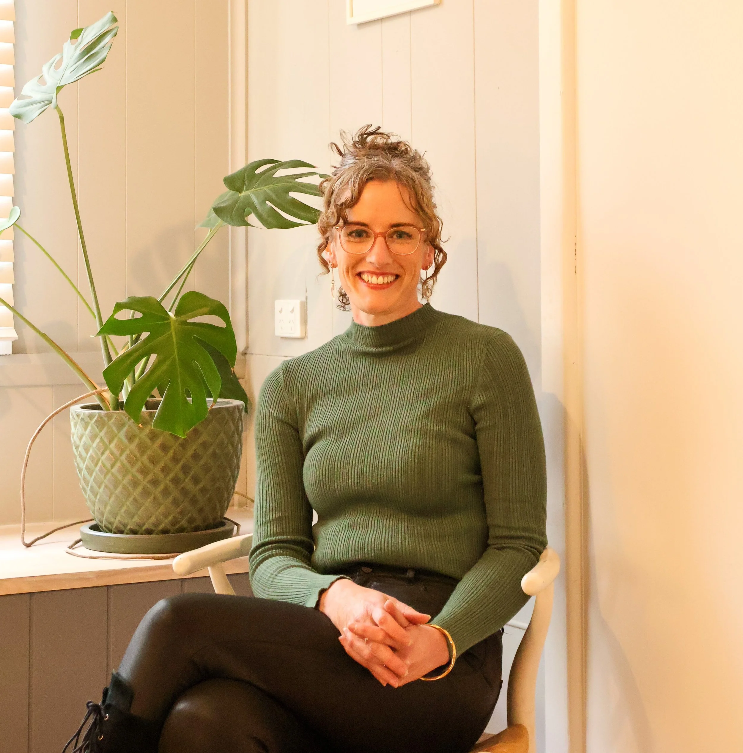 Woman in a red sleeveless top sitting on a couch, smiling, with indoor plant and window blinds in the background.