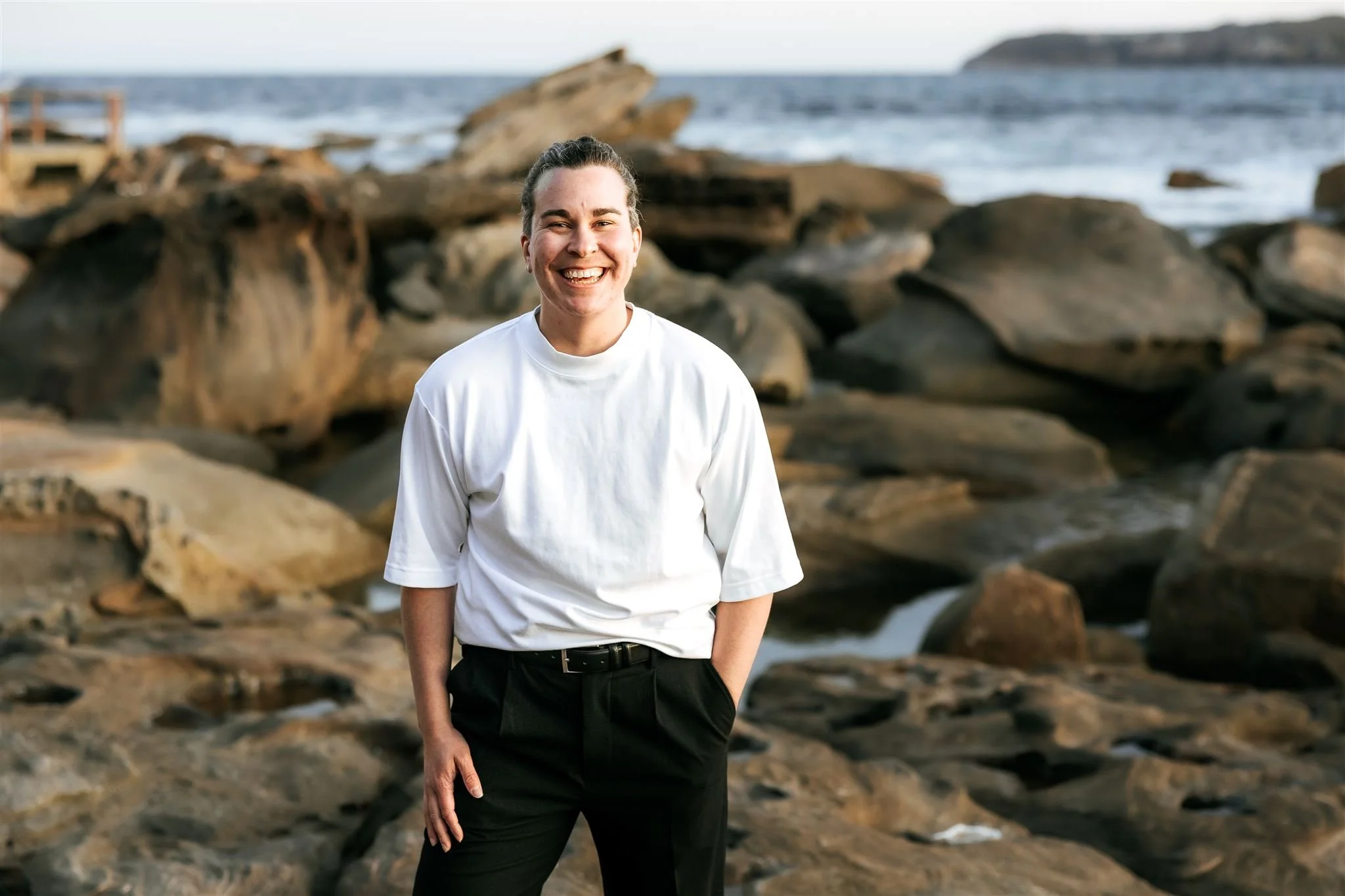 A person smiling and standing on a rocky beach with the ocean in the background. Steff Fenton