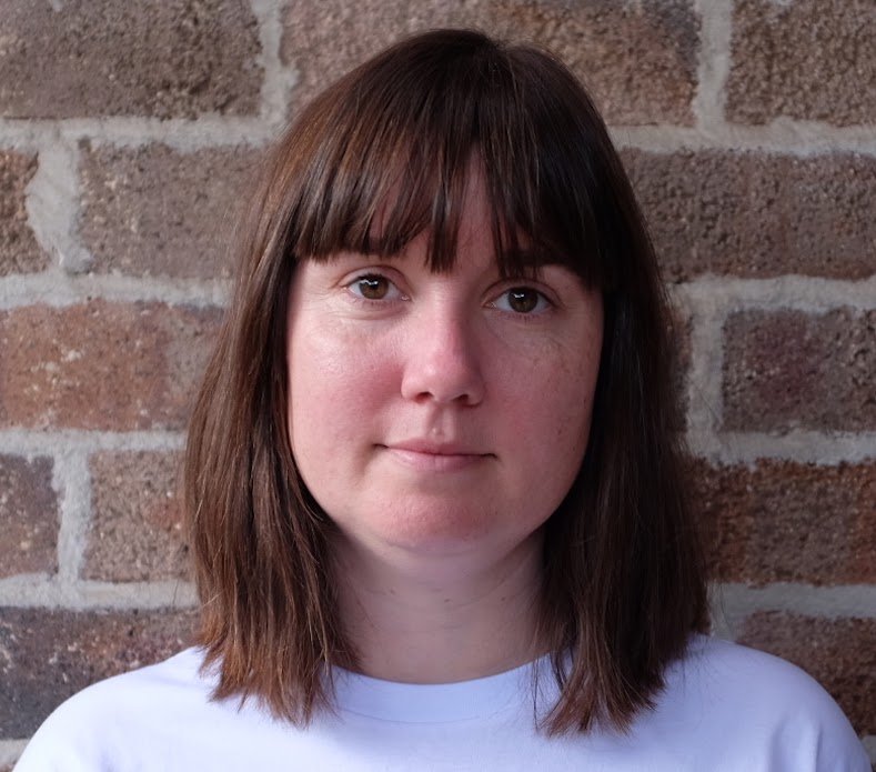 A woman with straight, shoulder-length brown hair and bangs standing in front of a brick wall, wearing a white top. Sarah Steel