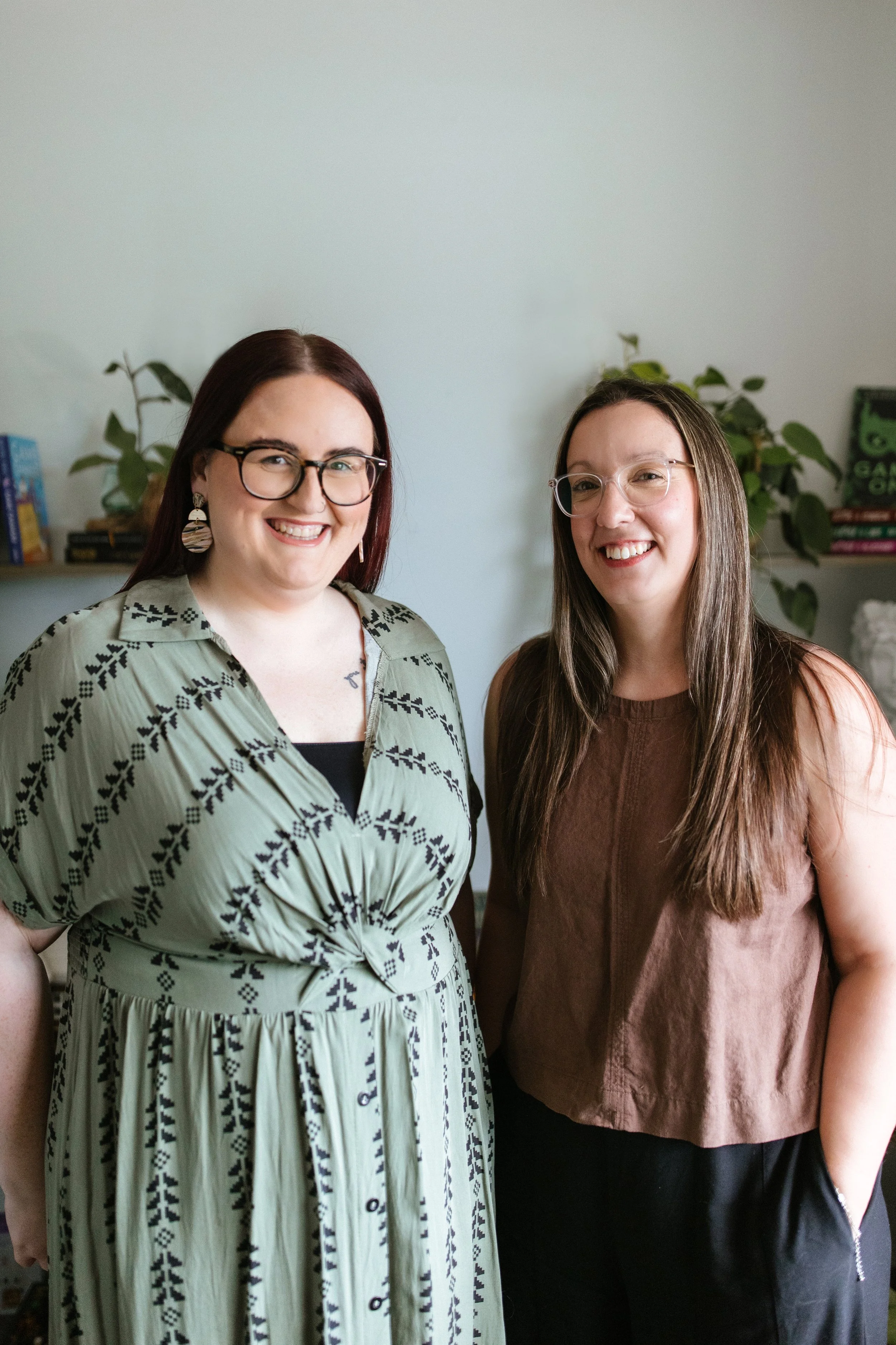 Two smiling women standing indoors in front of a light-colored wall with books and plants on shelves behind them. Elise Heerde & Sam Sellers