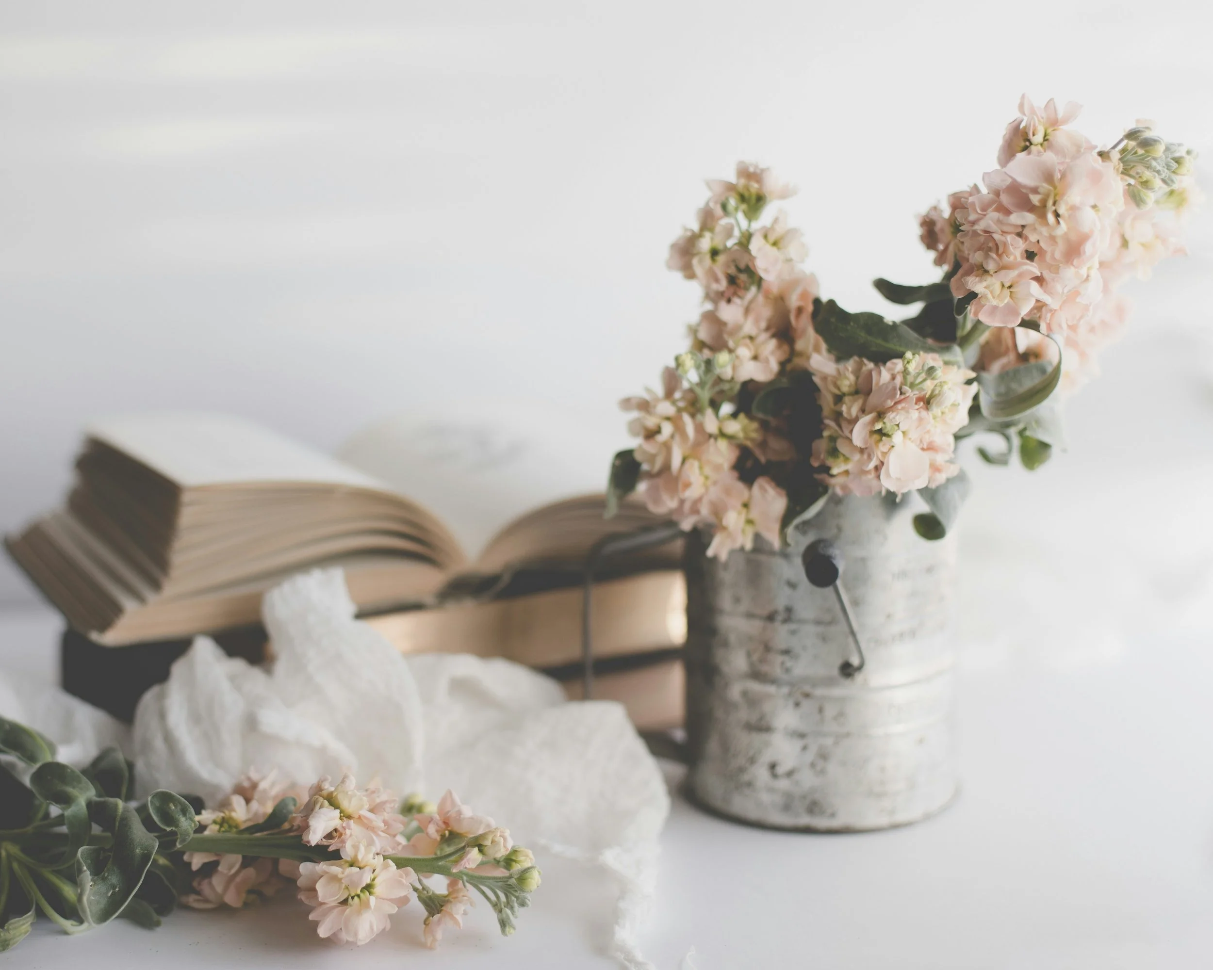 A bouquet of light pink and white flowers in a rustic metal container, with an open book and decorative rocks nearby on a white surface.