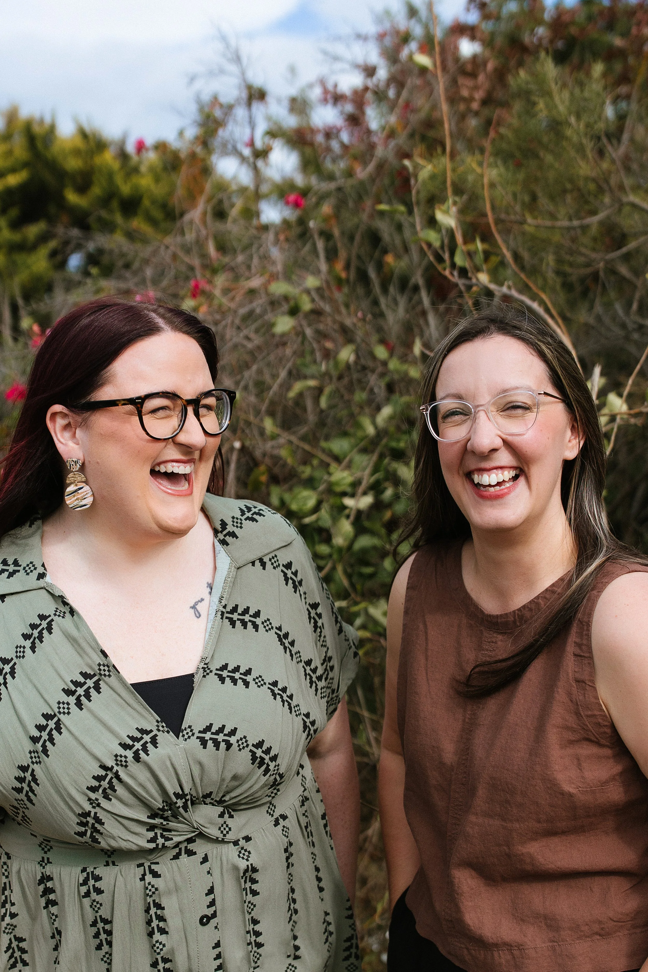 Two women smiling and laughing outdoors in front of a garden with green and red foliage. Elise Heerde & Sam Sellers