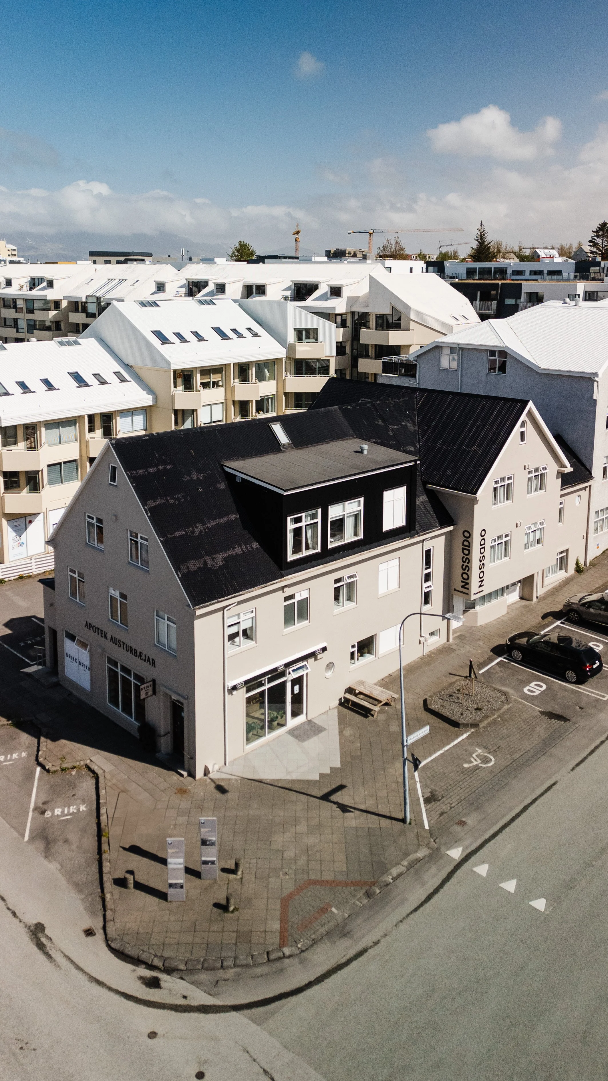 Aerial view of a city street showing a modern building with multiple signs and parking spots, surrounded by other urban buildings.