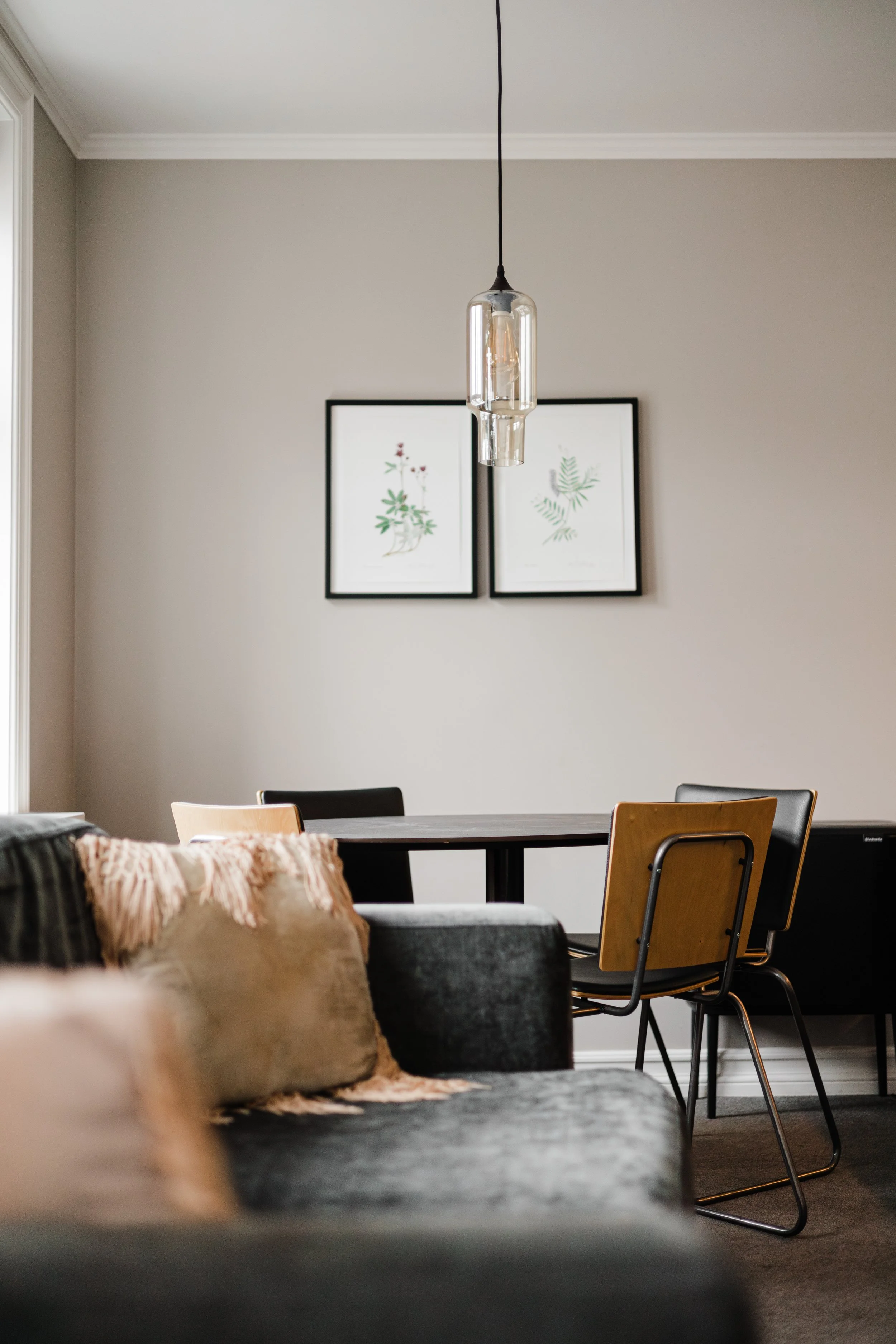 Living room with gray sofa, pillows, a dining table with black and wooden chairs, framed botanical prints on the wall, and a hanging pendant light.
