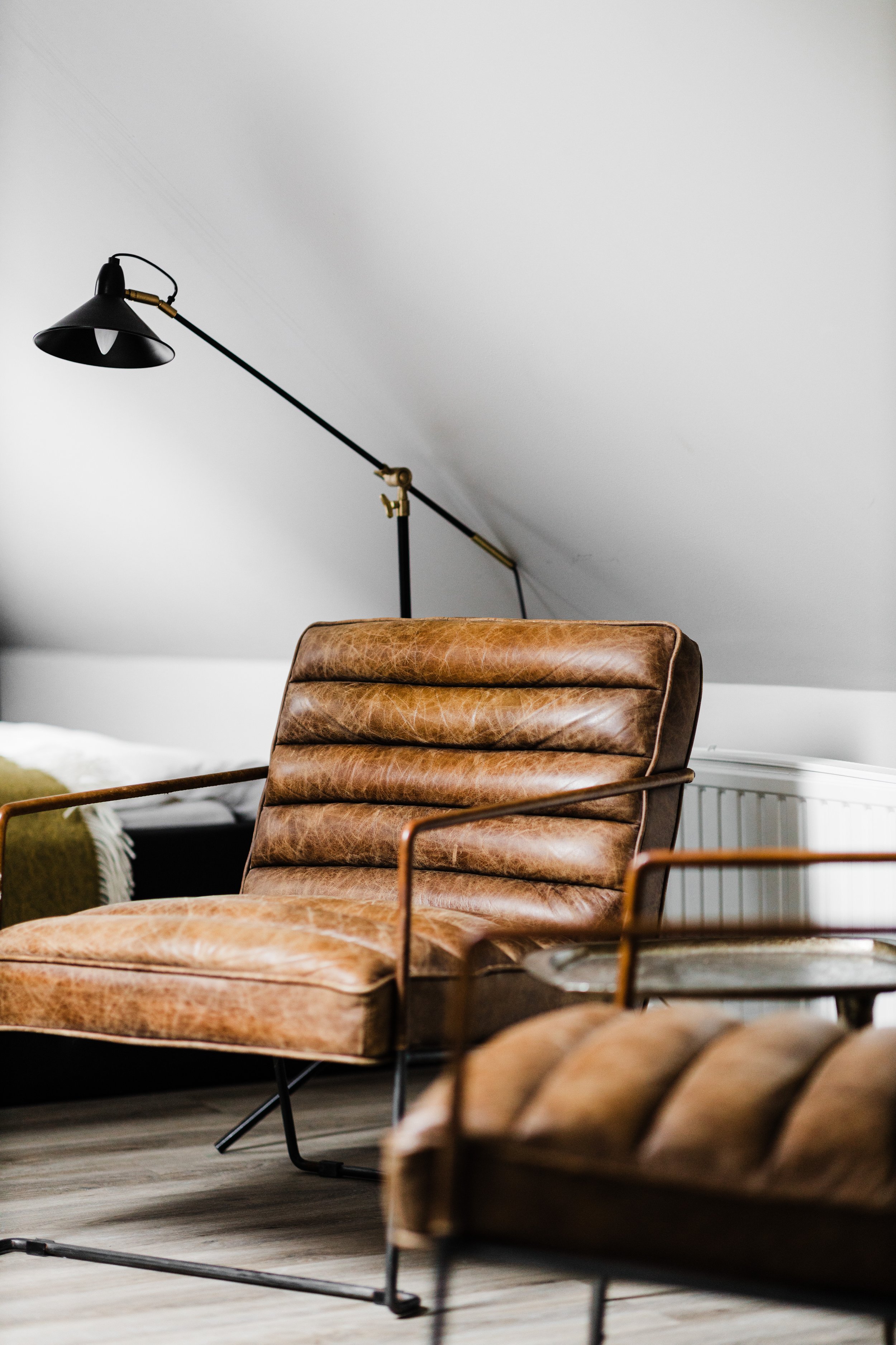 A vintage brown leather lounge chair with a metal frame in a modern indoor space, with a black adjustable wall-mounted lamp and a gray wall in the background.