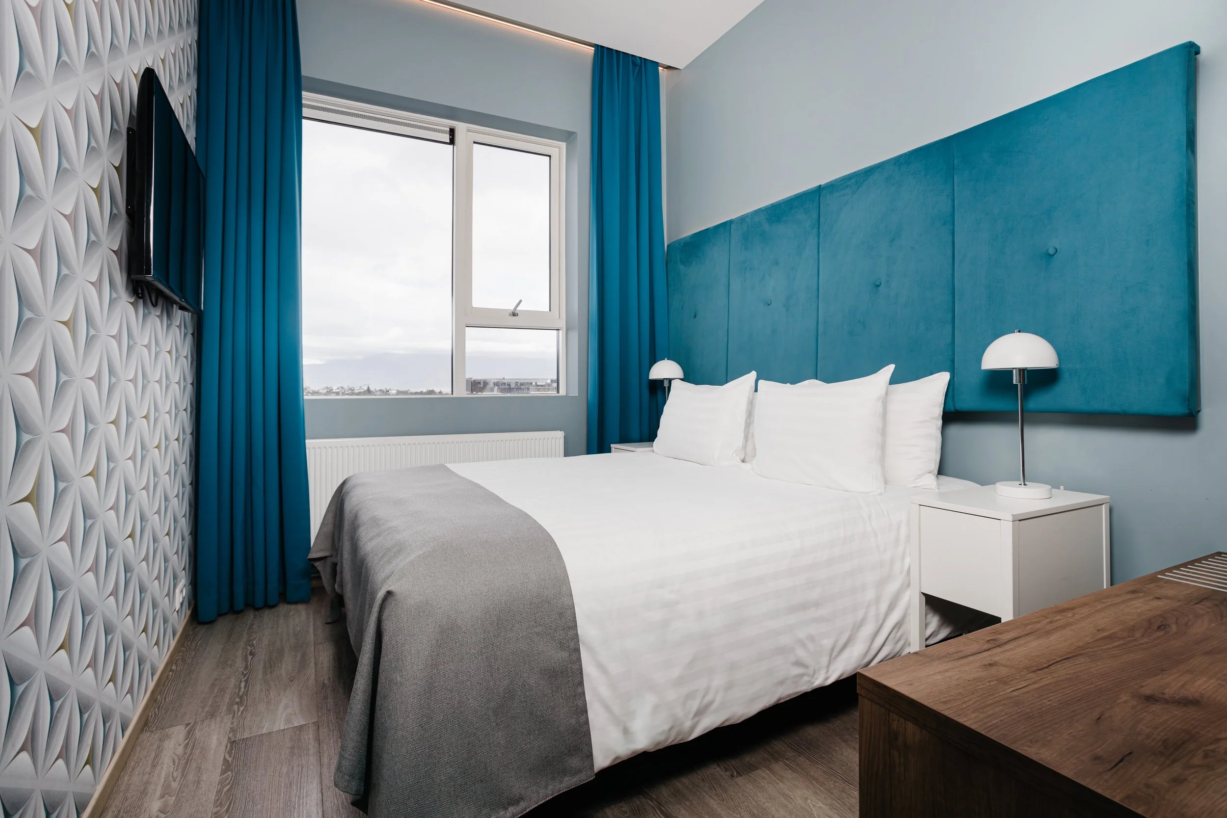 Hotel room with a bed, white linens, three pillows, gray throw blanket, blue headboard, white side table, lamp, window with blue curtains, patterned wall, and a TV mounted on the wall.