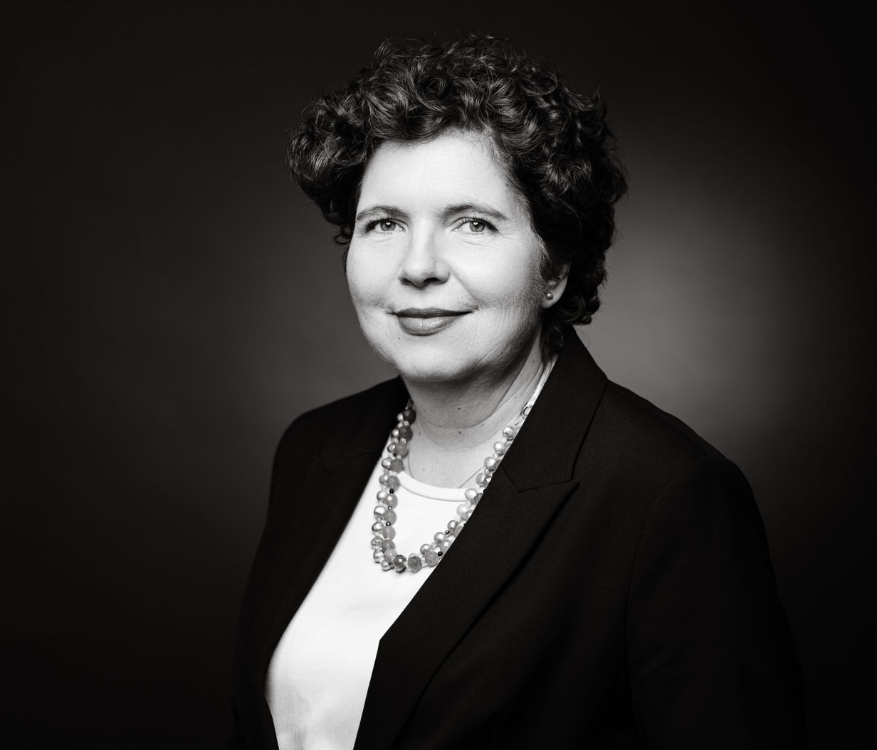Black and white professional portrait of a woman with curly hair, wearing a blazer, white blouse, and a beaded necklace, against a plain dark background.