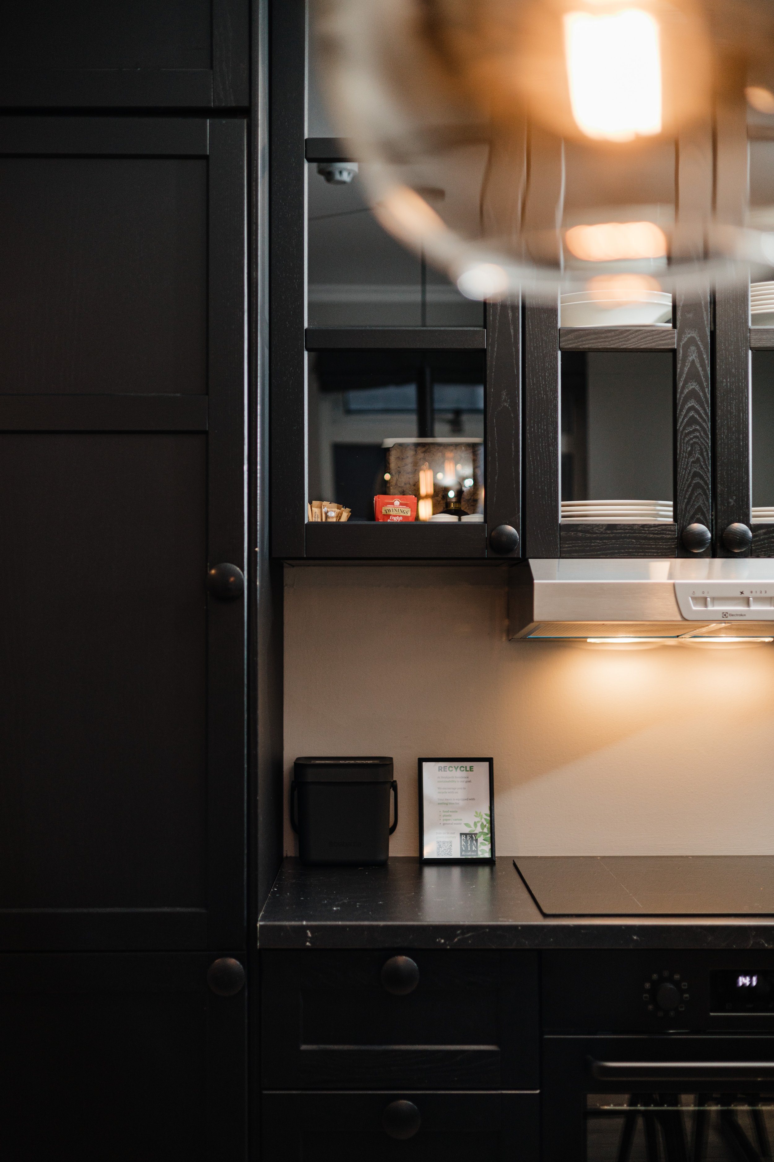 Close-up of a modern kitchen with black cabinets, a black countertop, and a beige wall, showing a small black container, a framed recycling info sign, and part of a stove.