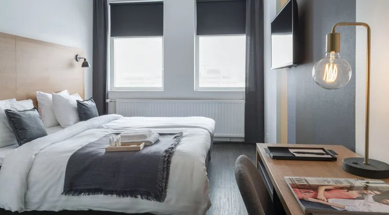 Modern hotel room with a neatly made bed, two windows with dark blinds, a desk with a magazine, and a desk lamp