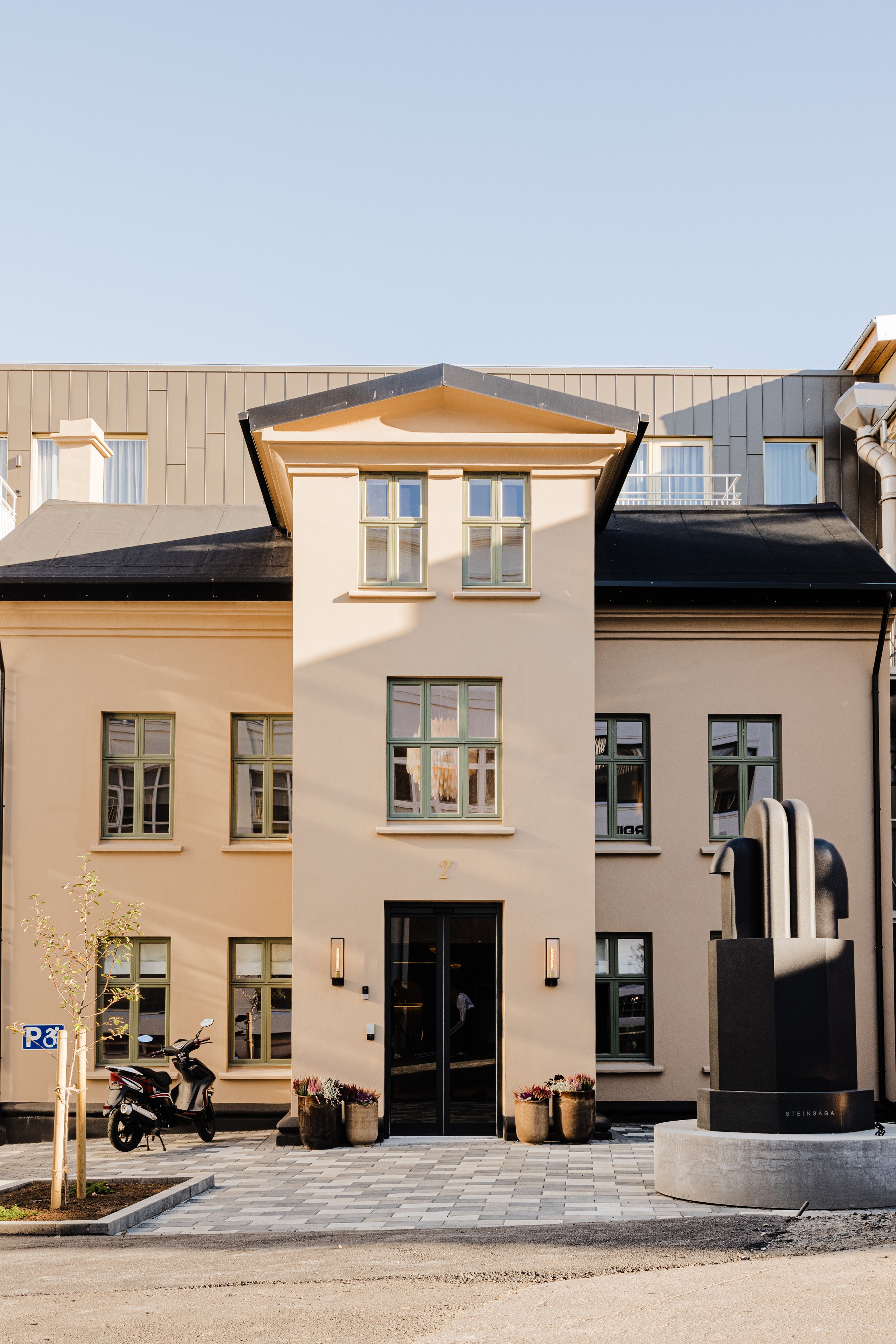 A modern residential building with beige walls, multiple windows, and a black front door. There are potted plants near the entrance, a parked scooter, and a large abstract sculpture to the right.