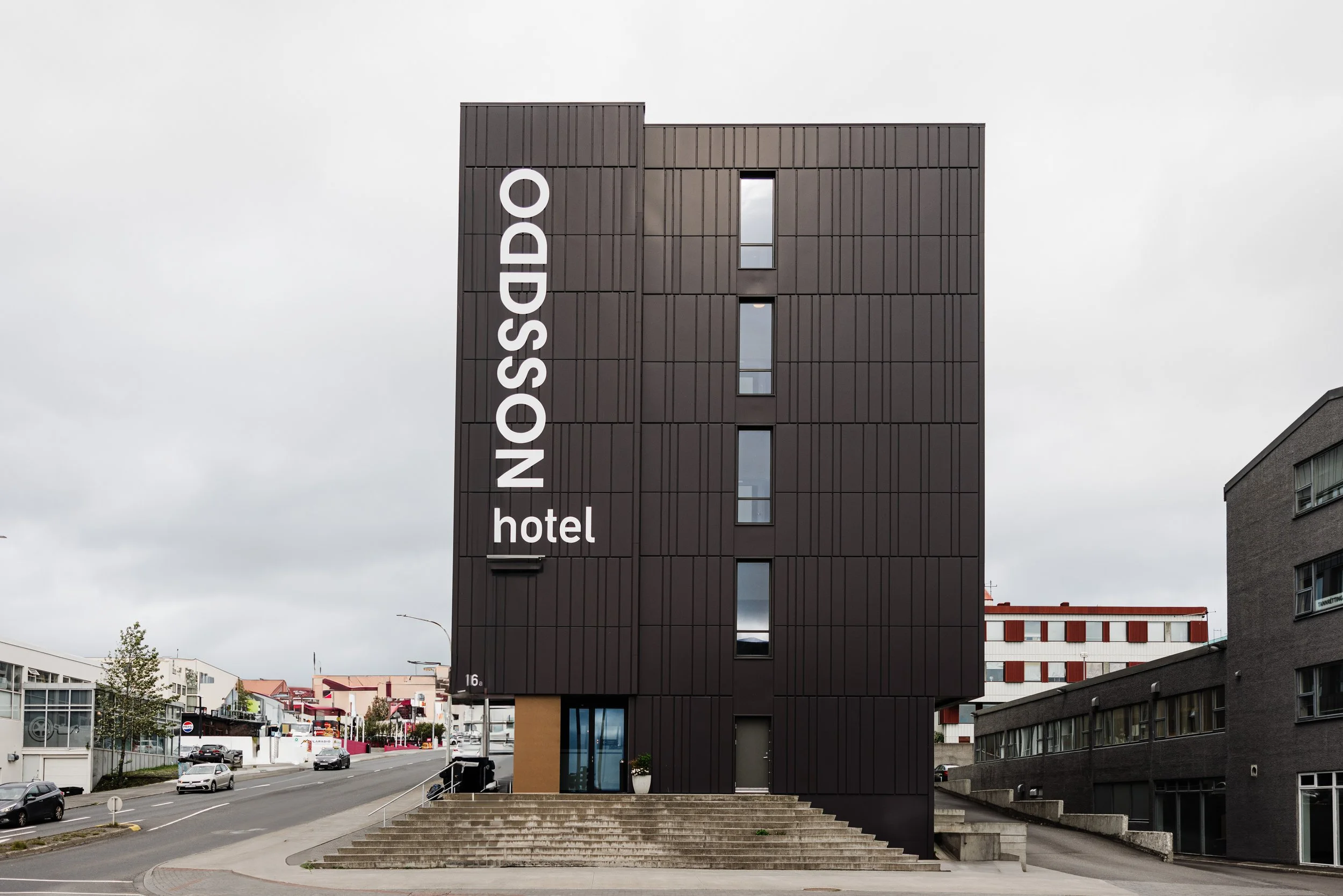 A modern black hotel building with the name 'ODDSSON hotel' in large white letters on the front. The building has narrow vertical windows and a set of steps leading to the entrance.