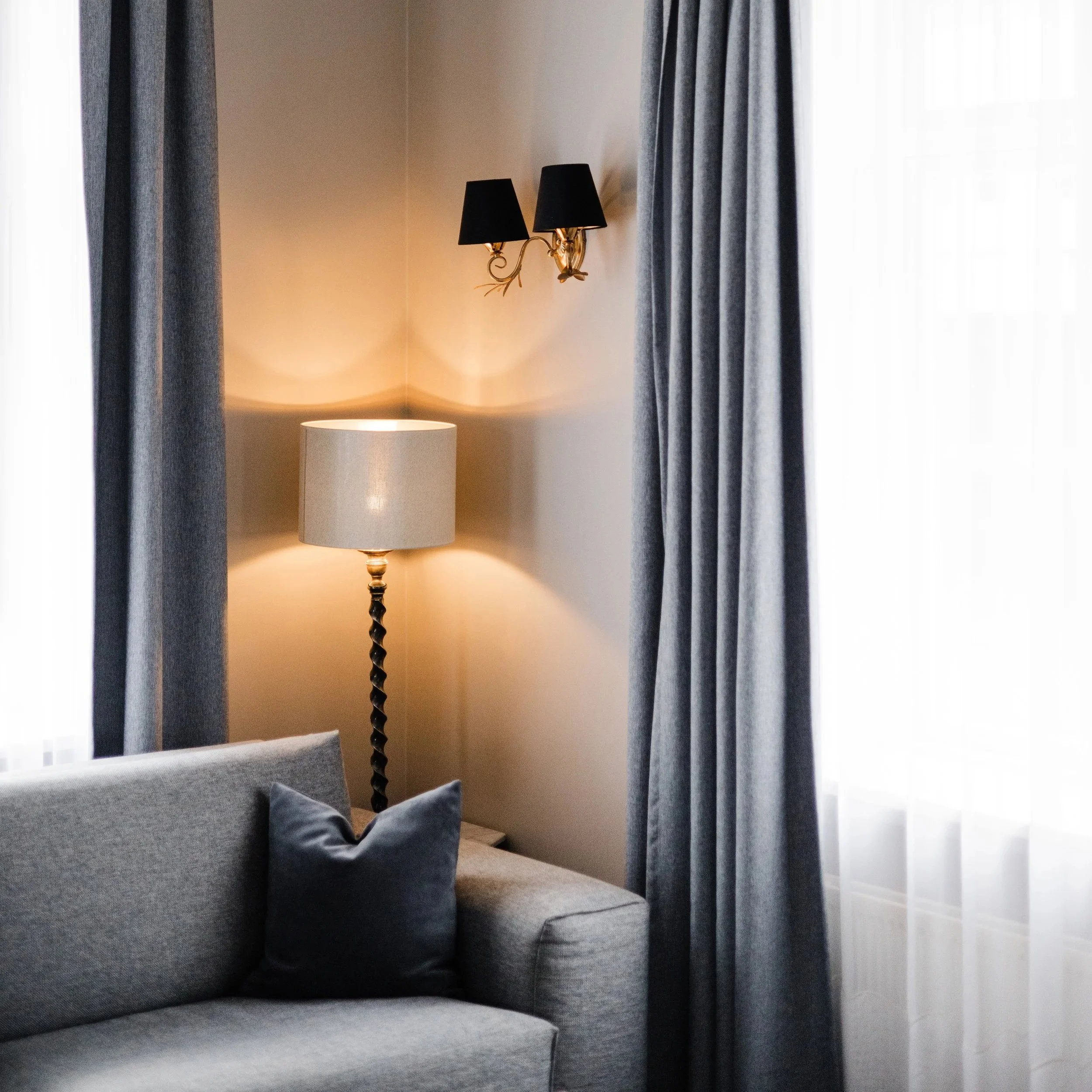Living room corner with gray sofa, gray curtains, a table lamp with a beige shade, and a wall-mounted black lampshade fixture.