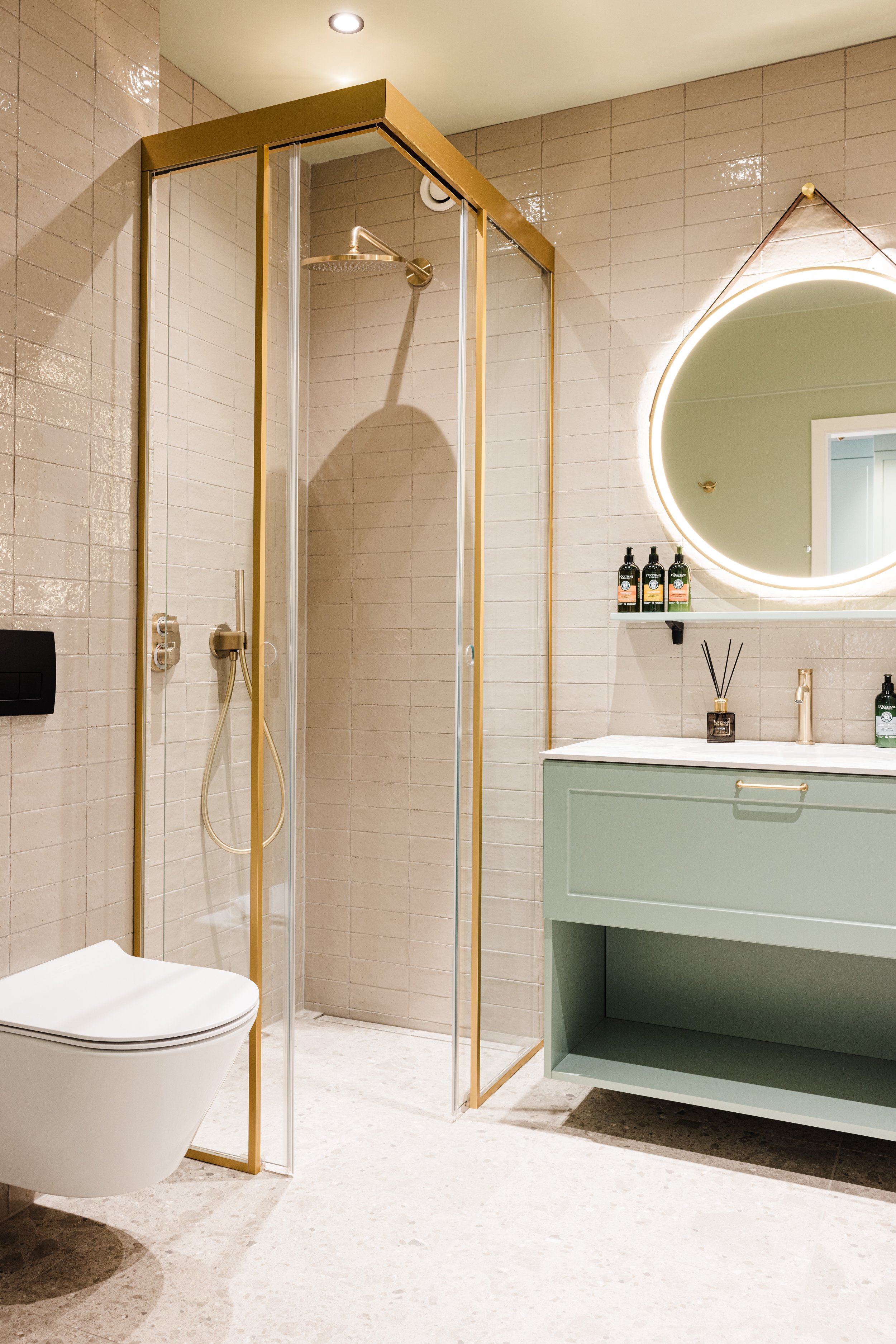 Modern bathroom with a glass shower enclosure with gold trim, a white wall-mounted toilet, and a mint green vanity with a large backlit round mirror above it. The vanity has a decorated shelf with bottles and a reed diffuser.
