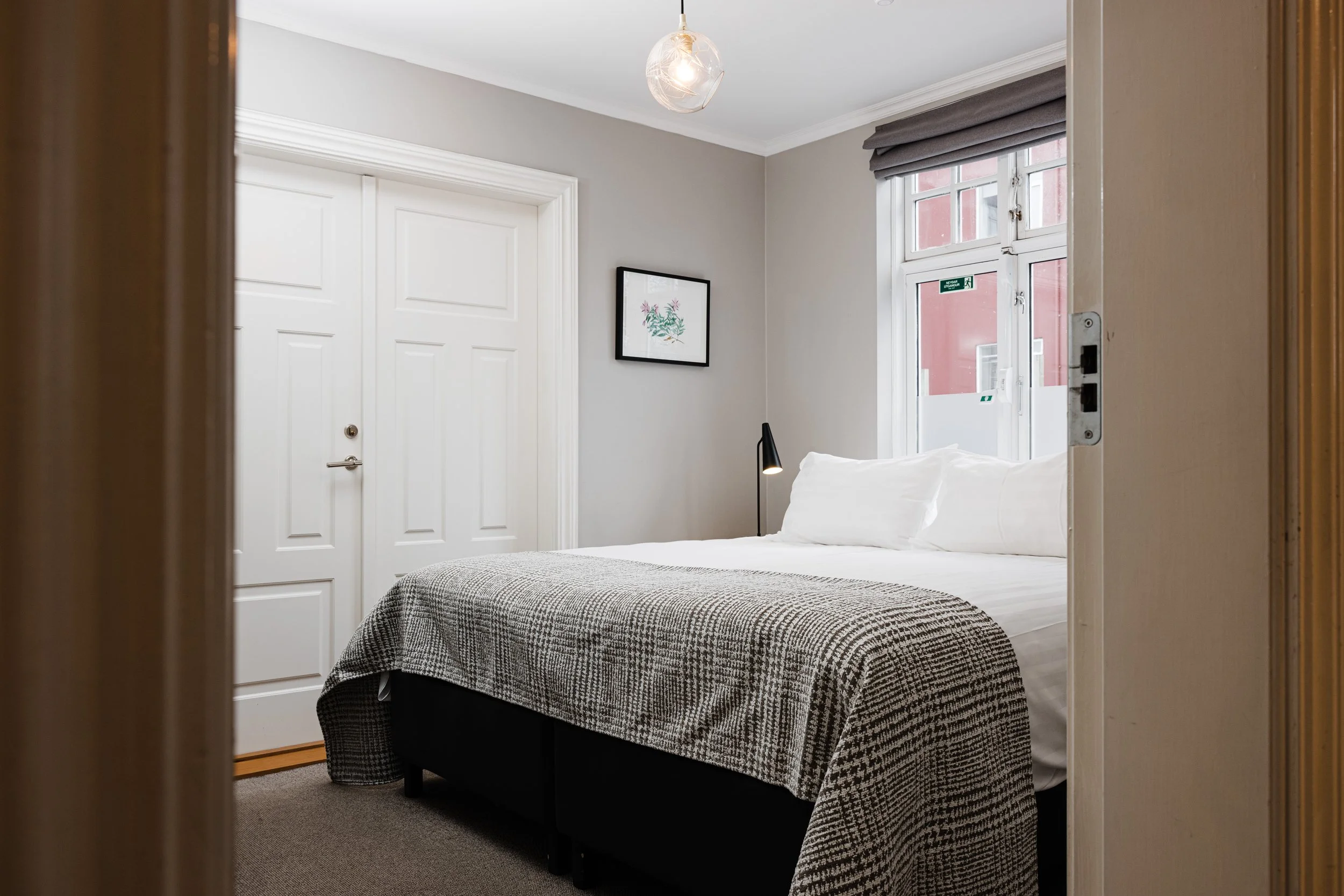 A cozy bedroom with a bed, white bedding, and pillows, next to a window with gray blinds, with a framed picture and a black bedside lamp.