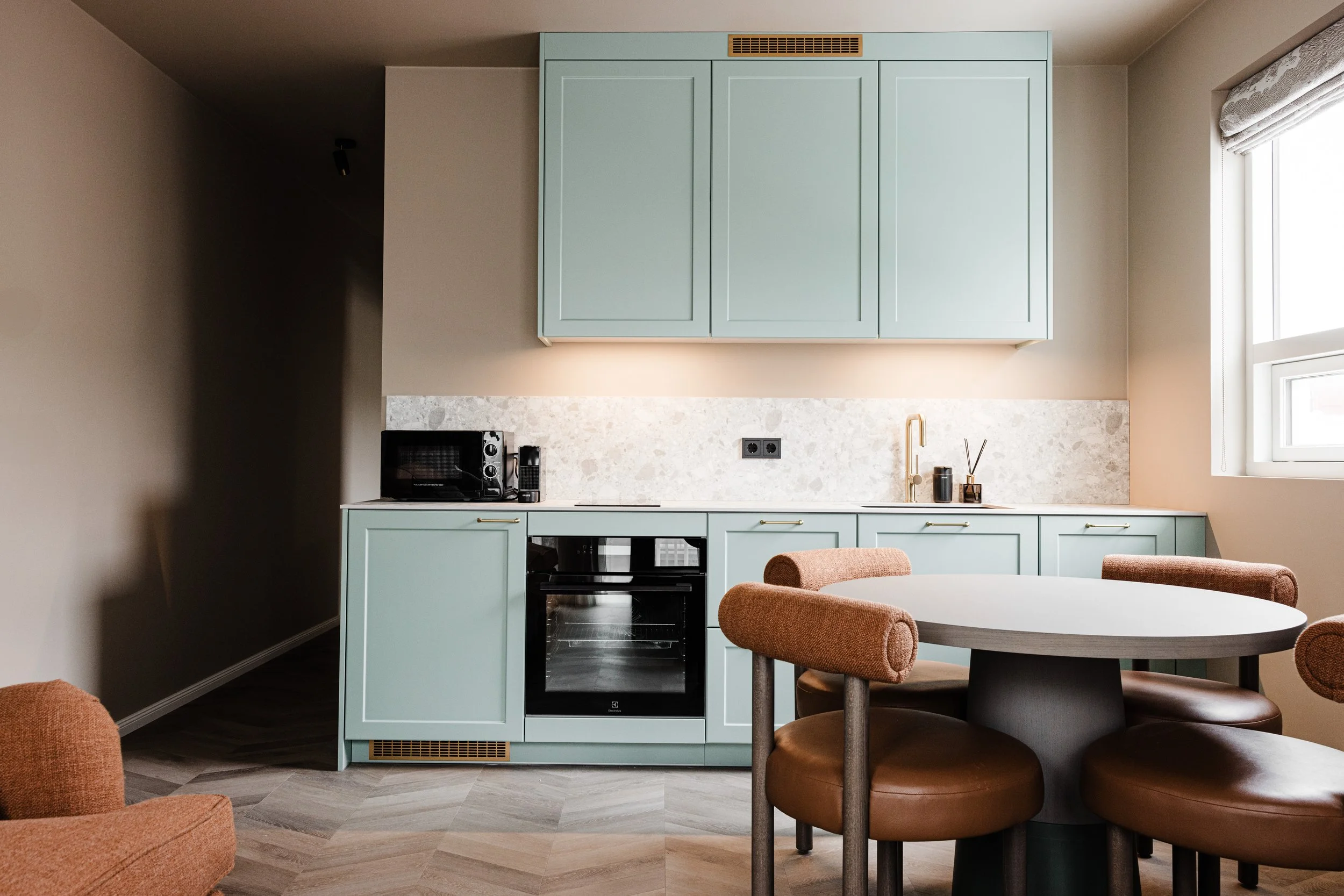 Modern kitchen with light green cabinets, a round dining table with four upholstered chairs, a window with a Roman shade, and small appliances on the counter.