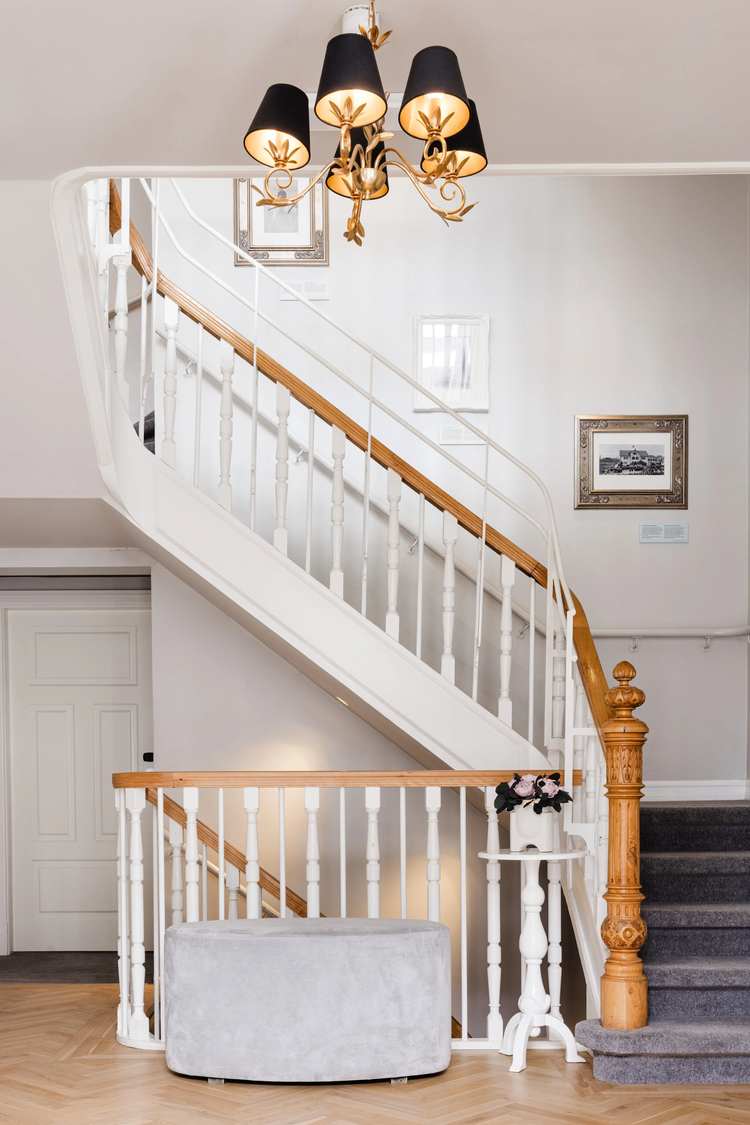 Interior view of a staircase with white and wood handrails, a gray carpeted staircase, a decorative chandelier, framed artwork on the wall, a white vase with pink flowers, and a gray upholstered bench.